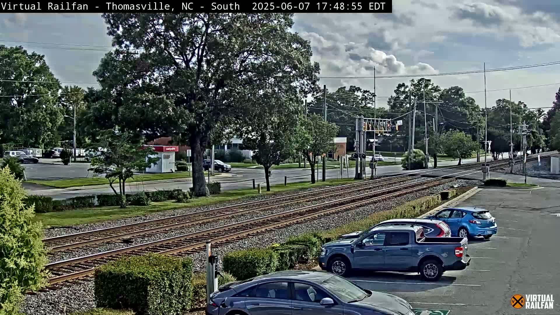 A partly cloudy day shows train tracks crossing a road in a suburban area with several cars parked nearby.