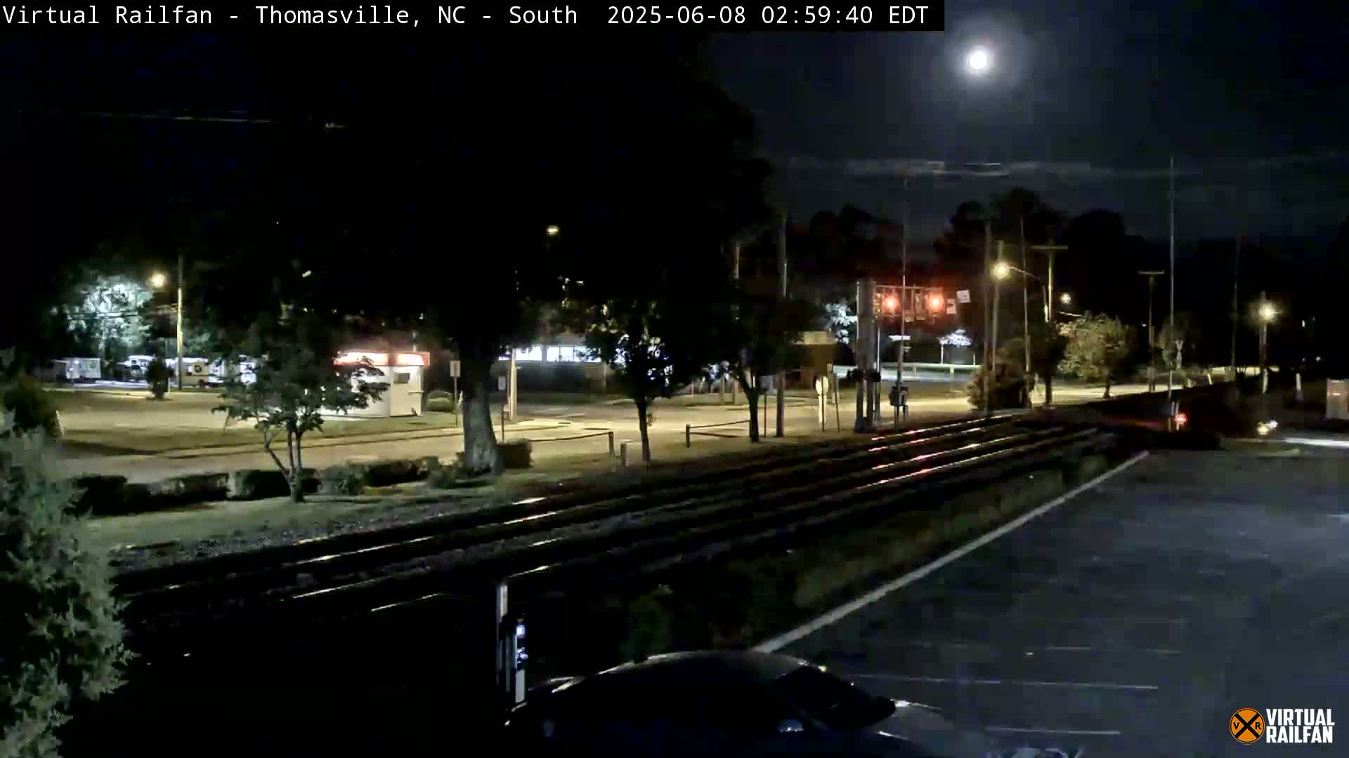 Under a bright moonlit night sky, railroad tracks run alongside a road with sparse nighttime traffic, lined with trees and buildings.