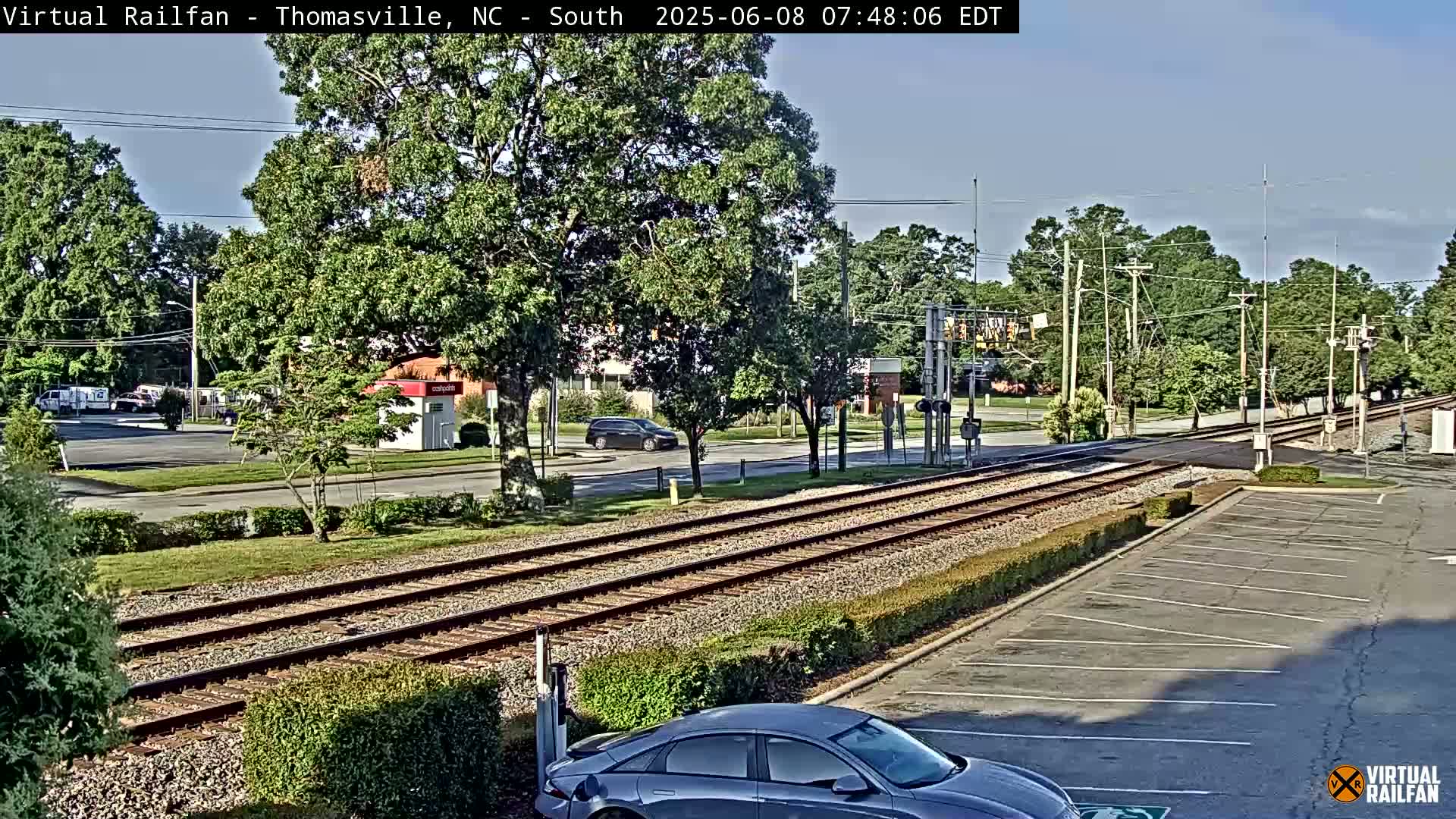 A view of railroad tracks crossing a road in a sunny area with lush green trees, cars parked and driving, and a small building in the distance.