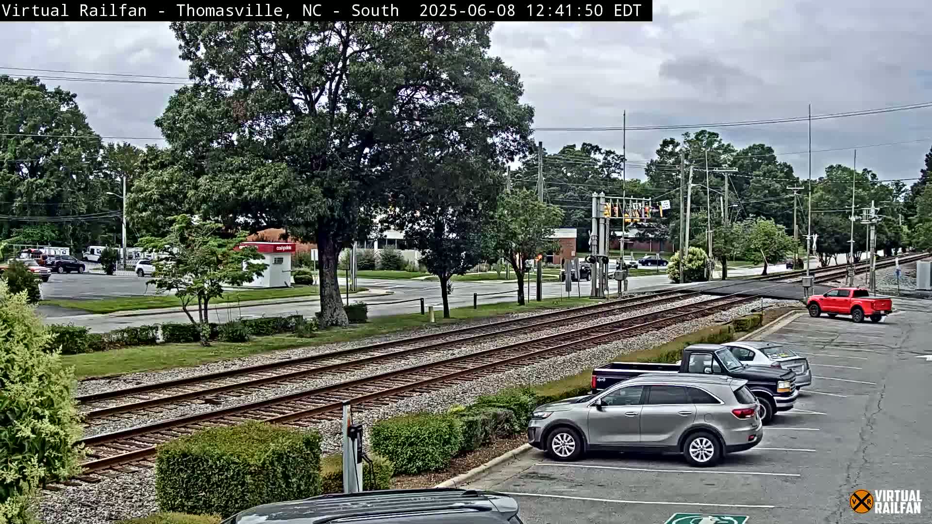 A view of railroad tracks intersecting a road, with several cars parked in a lot nearby, under a partly cloudy sky.