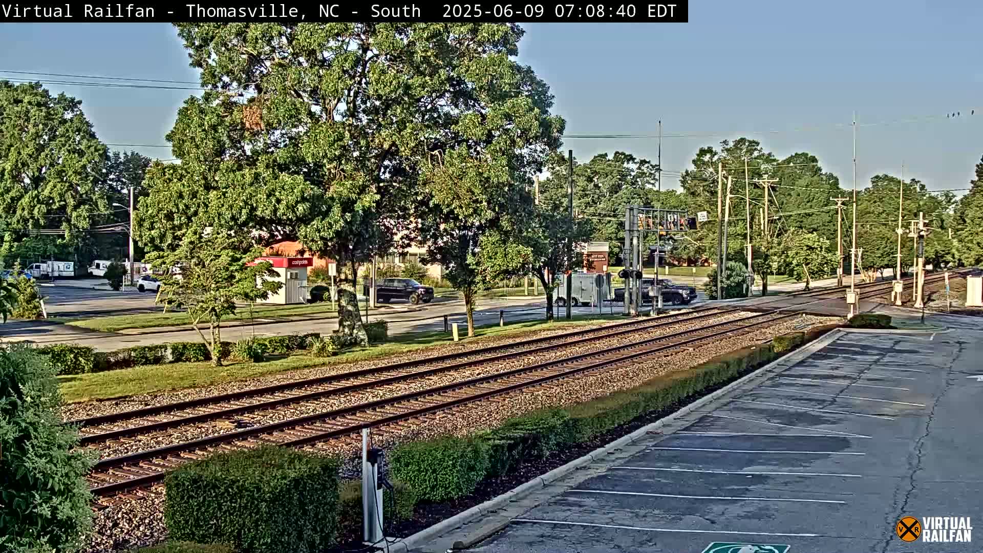 A sunny day shows a view of railroad tracks intersecting a road with cars and buildings in the background.
