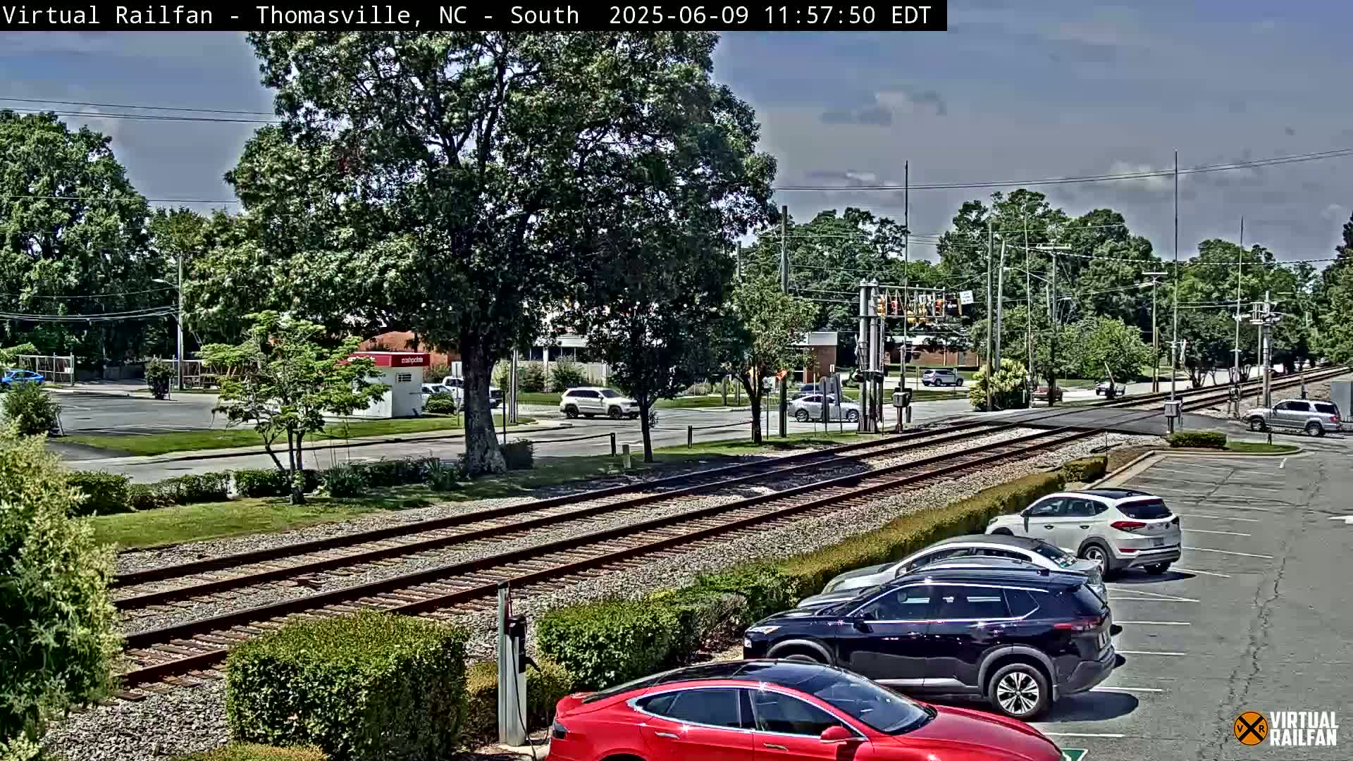 A partly sunny view of a street crossing train tracks, with several cars parked in a lot and others driving on the road.