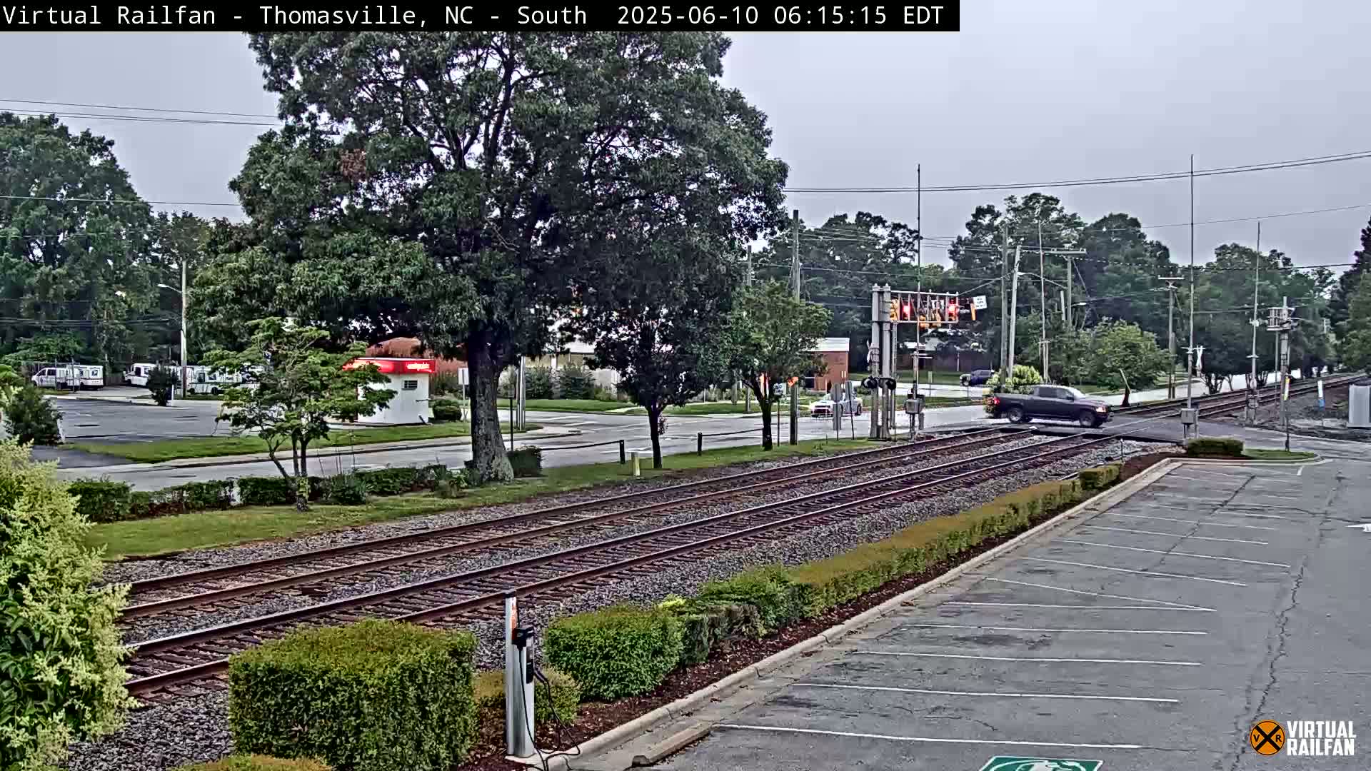 A dark-colored pickup truck crosses a railroad crossing under an overcast sky, with trees and buildings visible in the background.