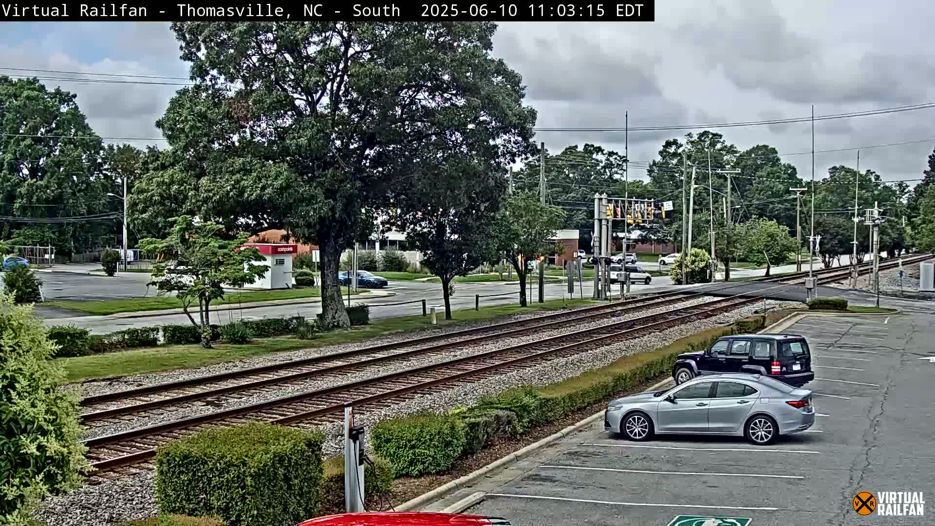Train tracks intersect a road in a partly cloudy setting, with several cars parked and driving nearby, surrounded by green trees and shrubs.