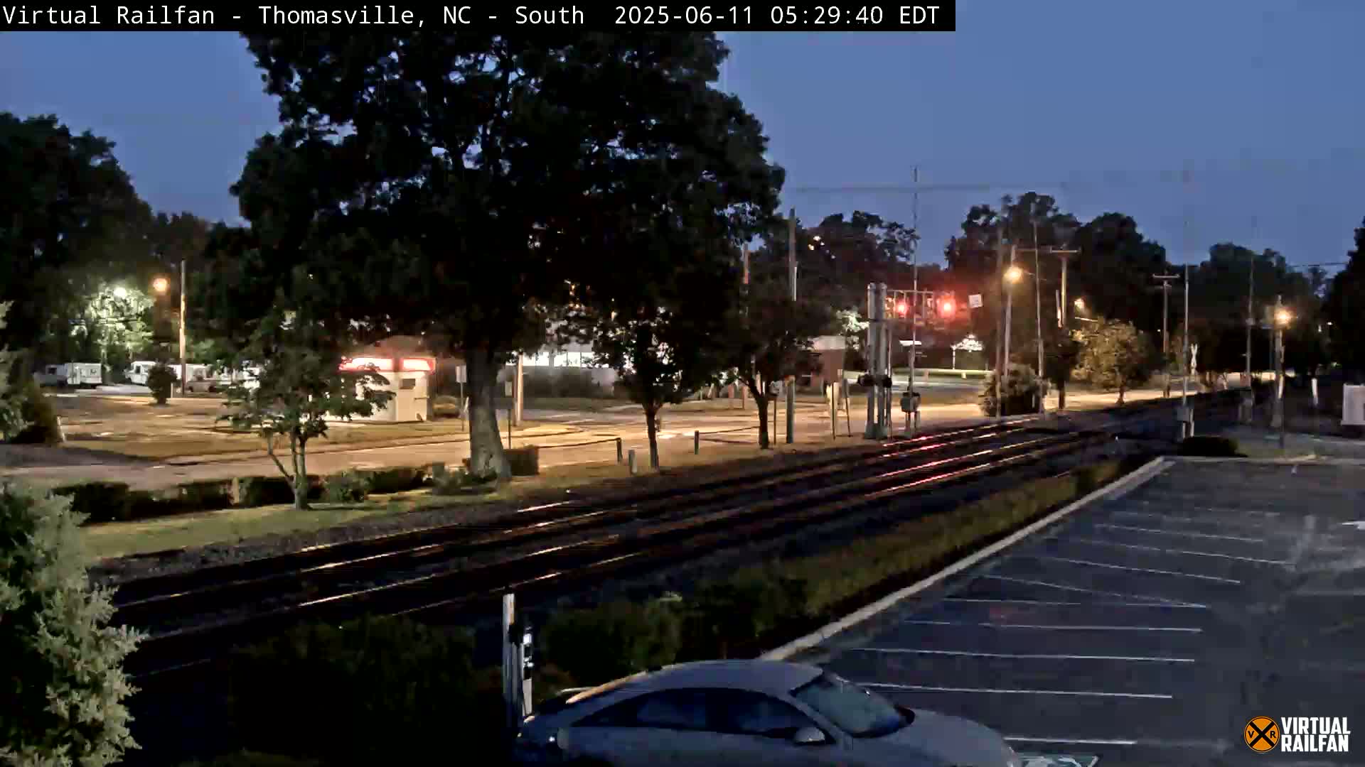 A twilight view shows railroad tracks running through a street intersection, lined with trees and streetlights, near a mostly empty parking lot with a car.