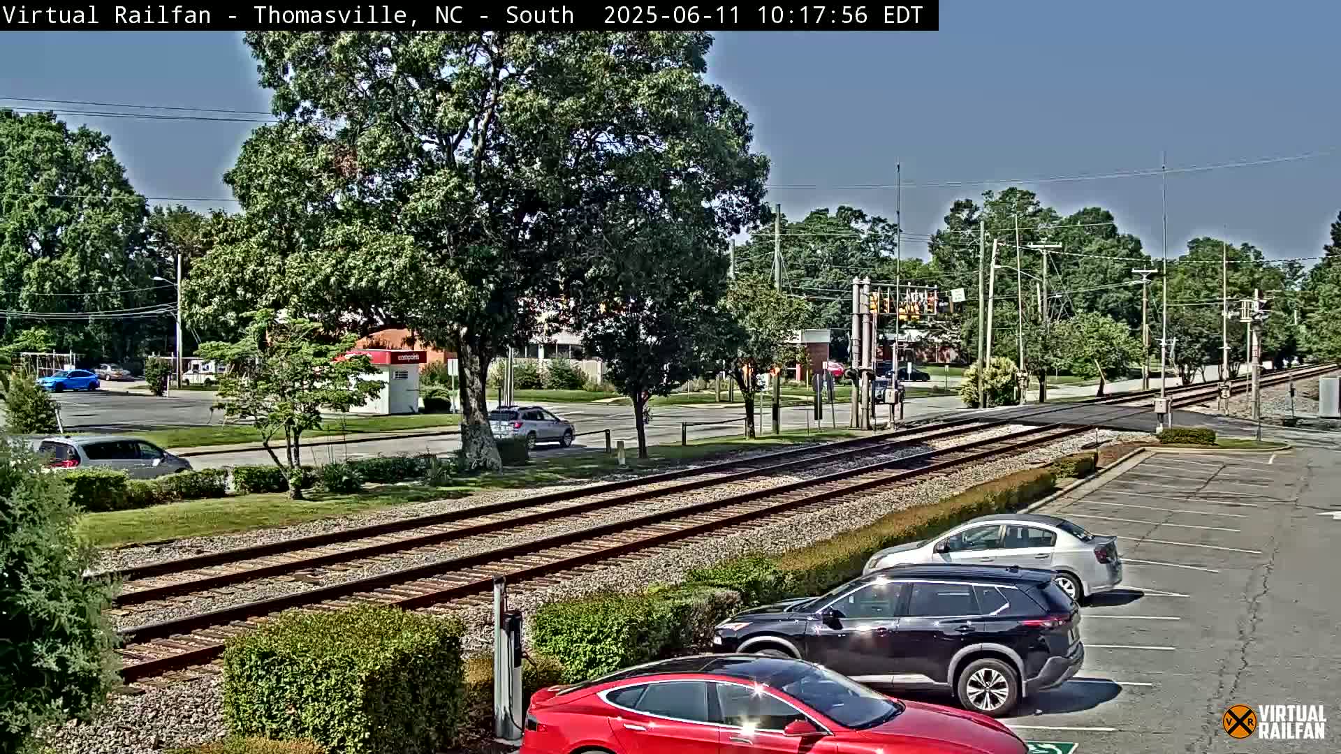 A sunny day shows a view of train tracks running through a town, with cars parked in a lot nearby and several vehicles driving on the road beside the tracks.