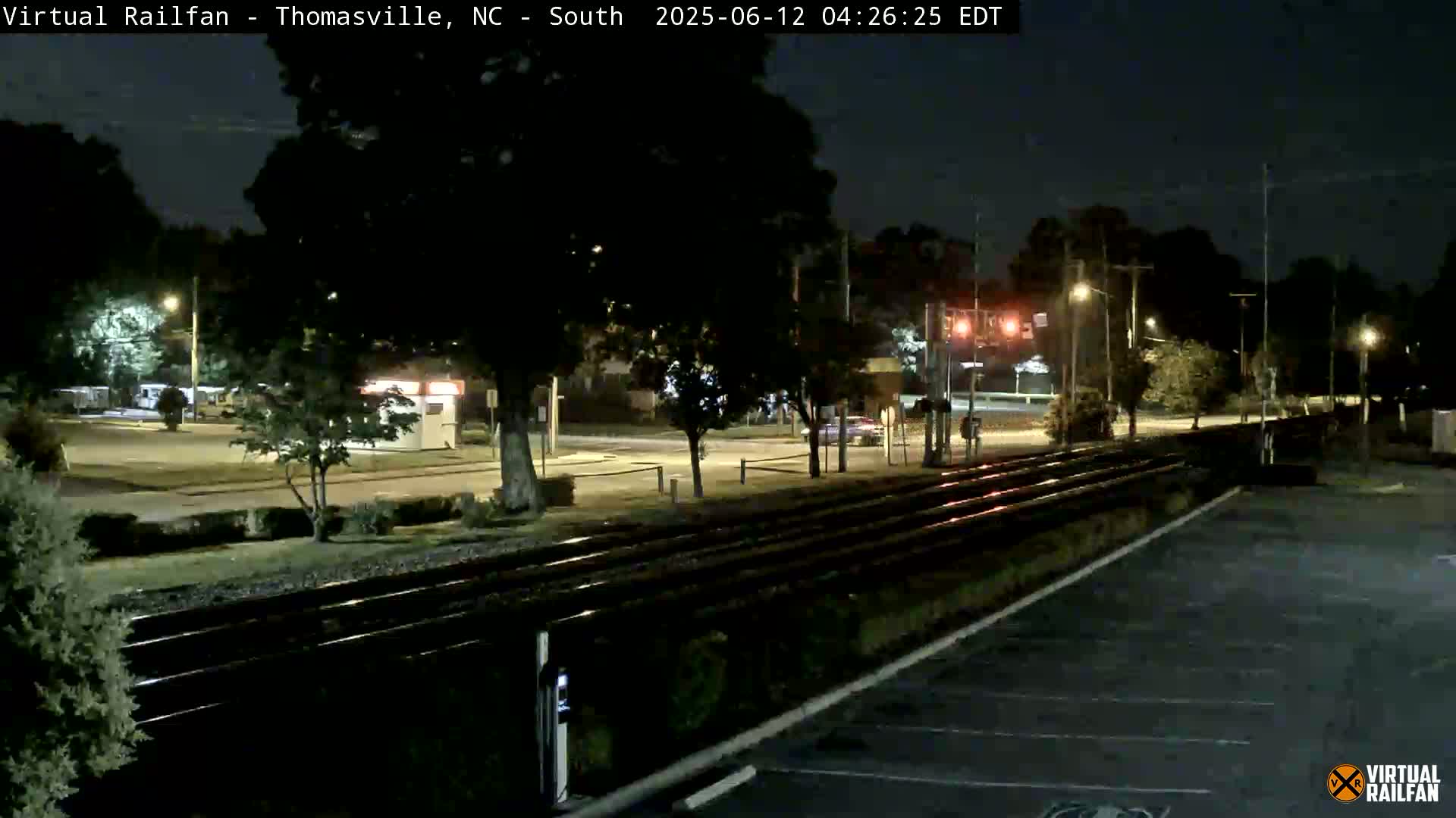 A nighttime view of railroad tracks next to a road with streetlights, trees, and a small building, under clear skies.