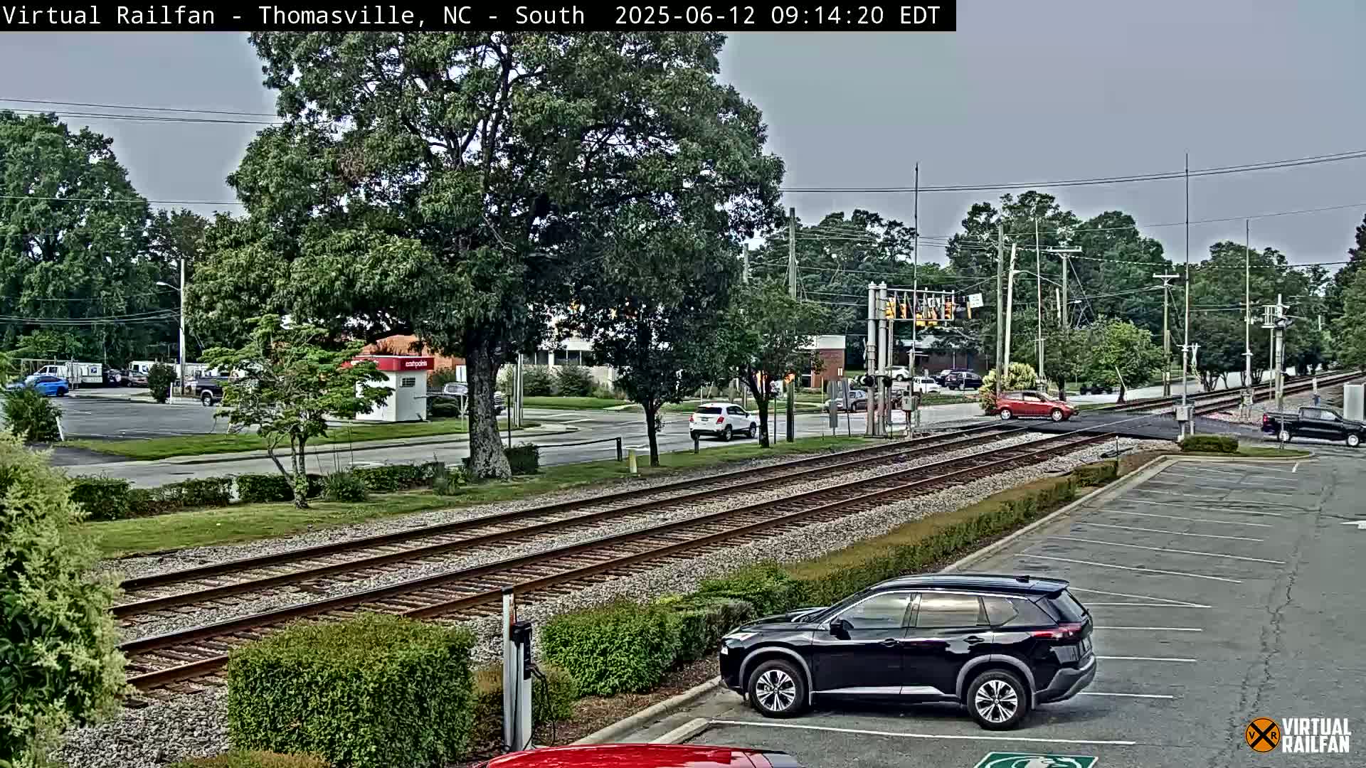 A view of a train crossing in a town on an overcast day, showing several cars parked and driving near railroad tracks.