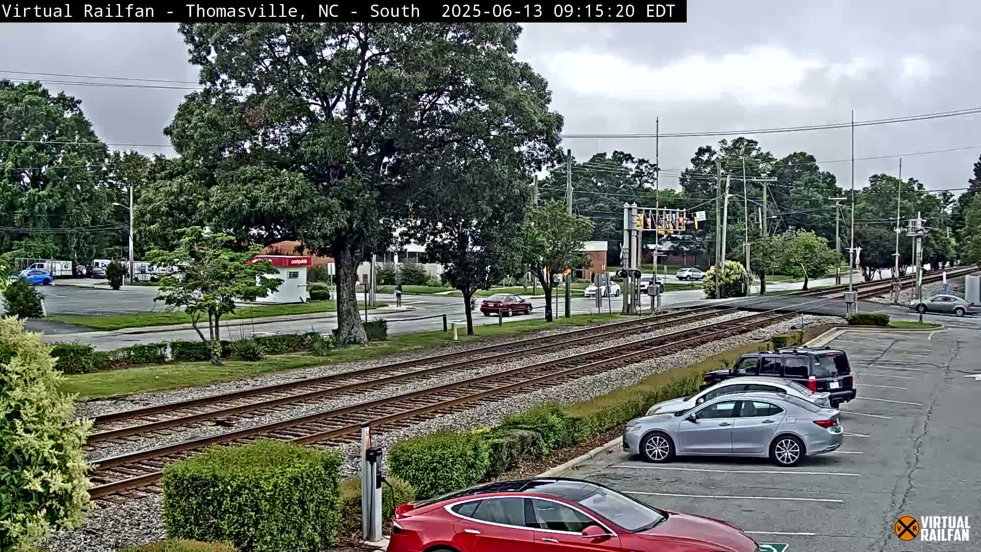A view of a railroad crossing in a town with overcast skies, showing cars parked in a lot beside the tracks and vehicles driving on the road.