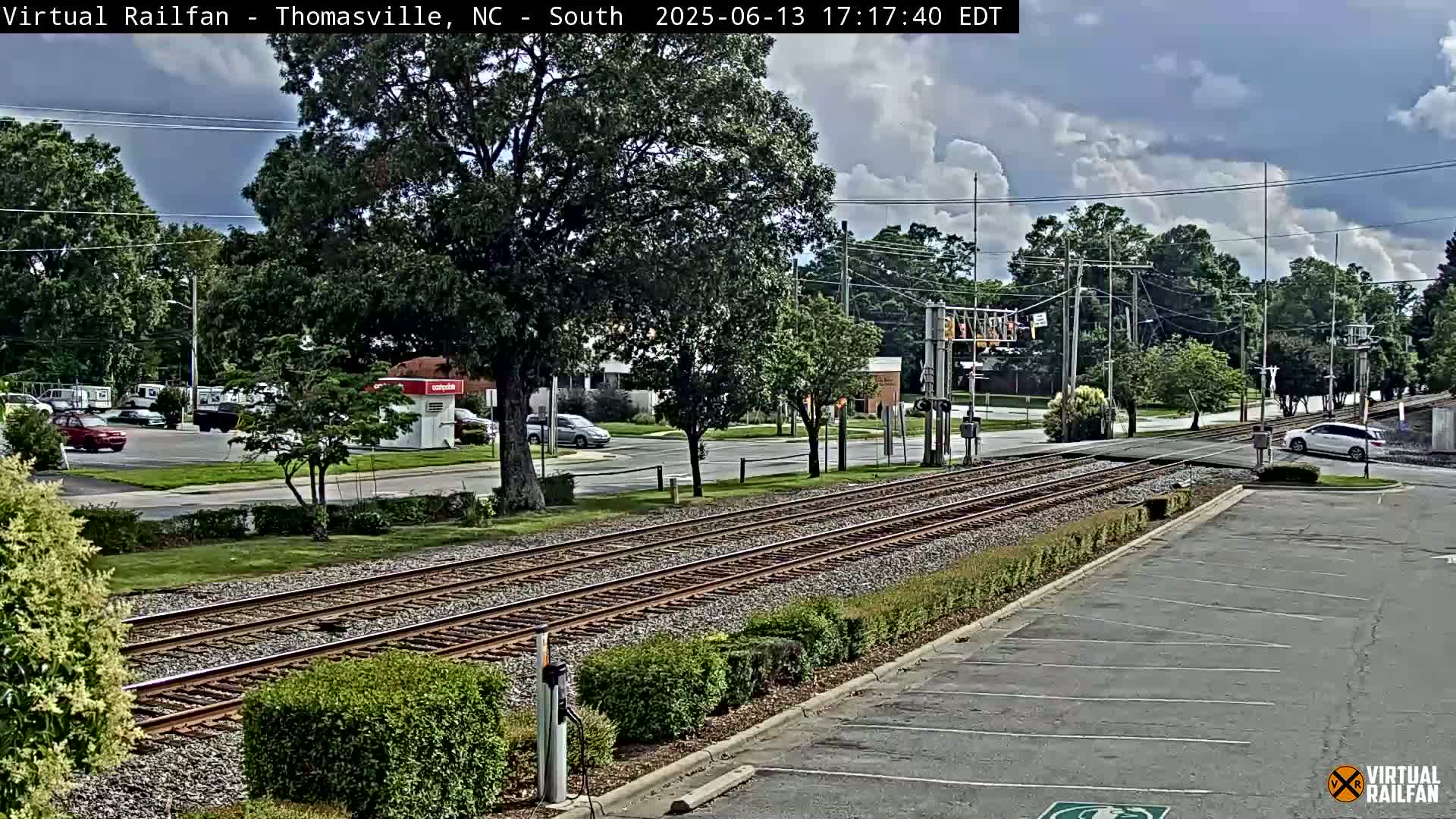 A partly cloudy day reveals train tracks intersecting a road lined with green shrubbery and trees, with several cars parked nearby.