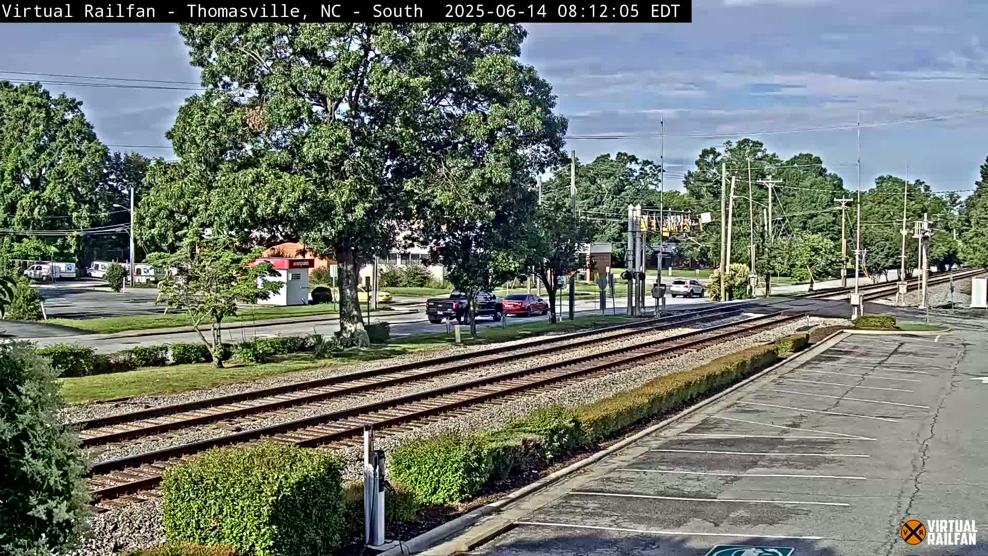 A partly sunny view of railroad tracks crossing a road, with several vehicles visible and a parking lot to the right, surrounded by lush green trees and shrubbery.