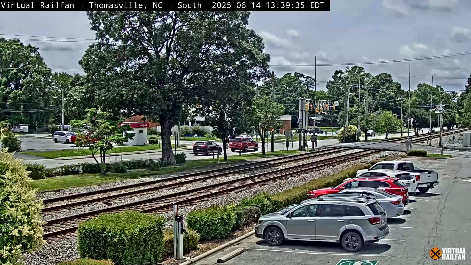 A partly cloudy day shows a view of railroad tracks intersecting a road with several cars parked in a lot and a few driving on the road.
