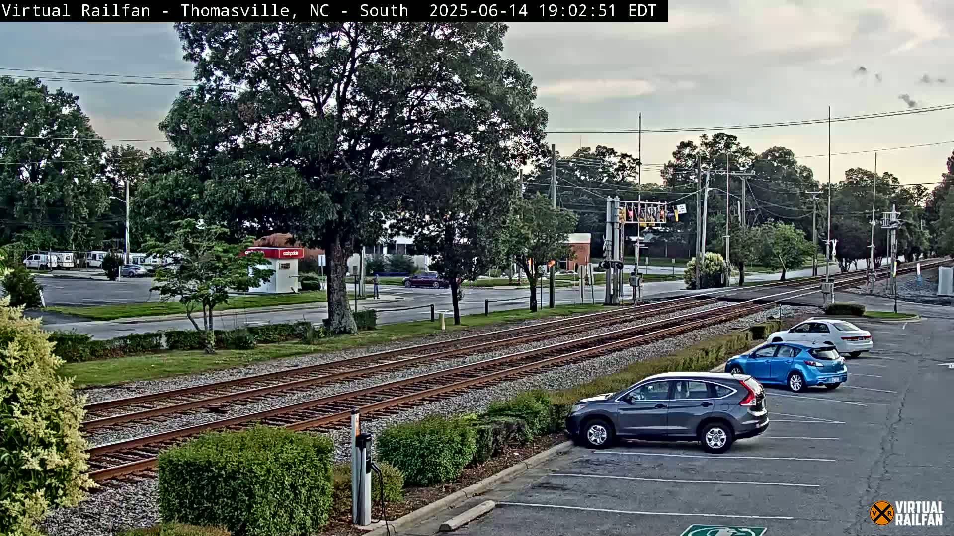 A partly cloudy day shows a view of railroad tracks intersecting a road with several cars parked in a lot and a few driving on the road.