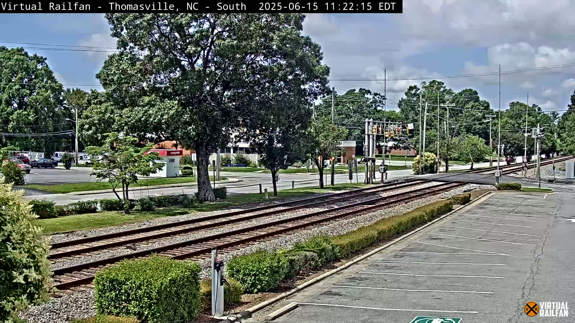 A partly cloudy day reveals a railroad crossing intersecting a street lined with trees and a parking lot, with several cars visible in a nearby lot.