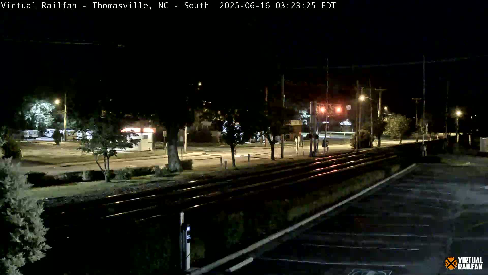 A nighttime view of train tracks intersecting a street, with streetlights illuminating a mostly empty parking lot and surrounding area.