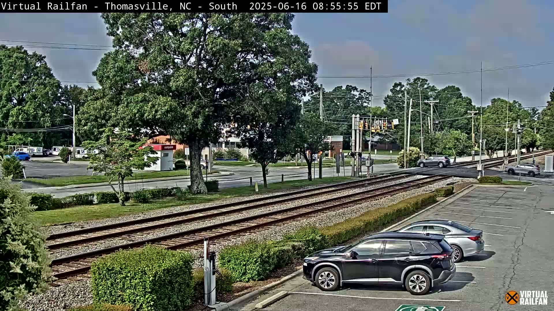 A mostly sunny day shows railroad tracks intersecting a road with several cars parked nearby, and one car crossing the tracks.