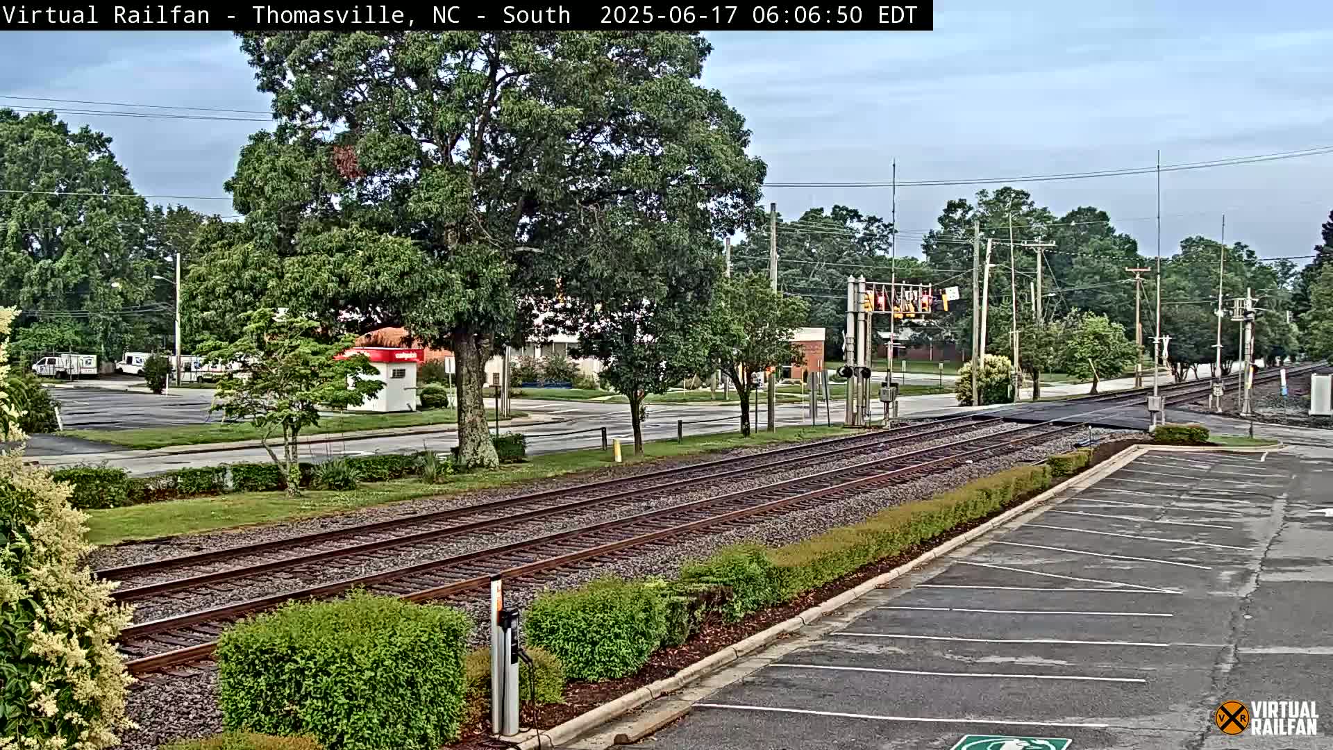 Train tracks intersect a road in a town on an overcast day, with lush green trees and shrubs lining the railway and parking lots nearby.