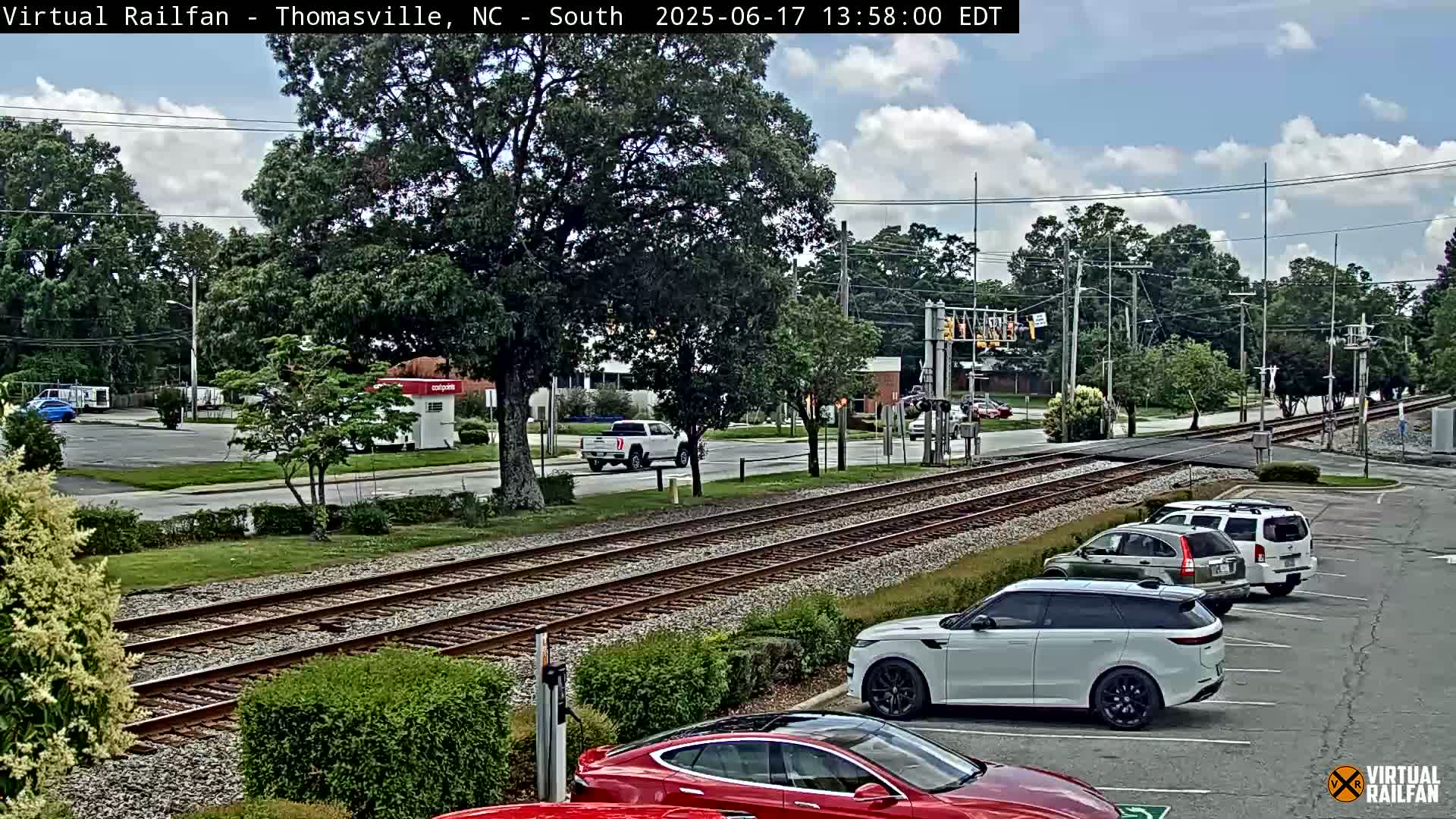 A partly cloudy day shows a view of railroad tracks intersecting a road with several cars parked in a lot and driving on the road, and trees lining the sides of the road.