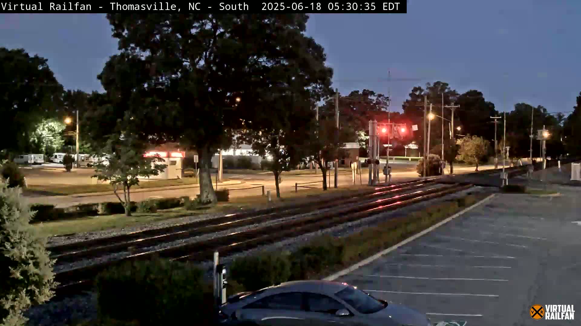 A twilight view of train tracks at a road crossing, with a car parked nearby and trees lining the area.