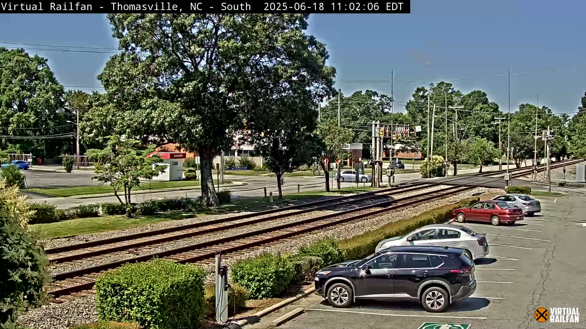 A sunny day view of railroad tracks intersecting a road, with several cars parked in a lot nearby and lush green trees in the background.