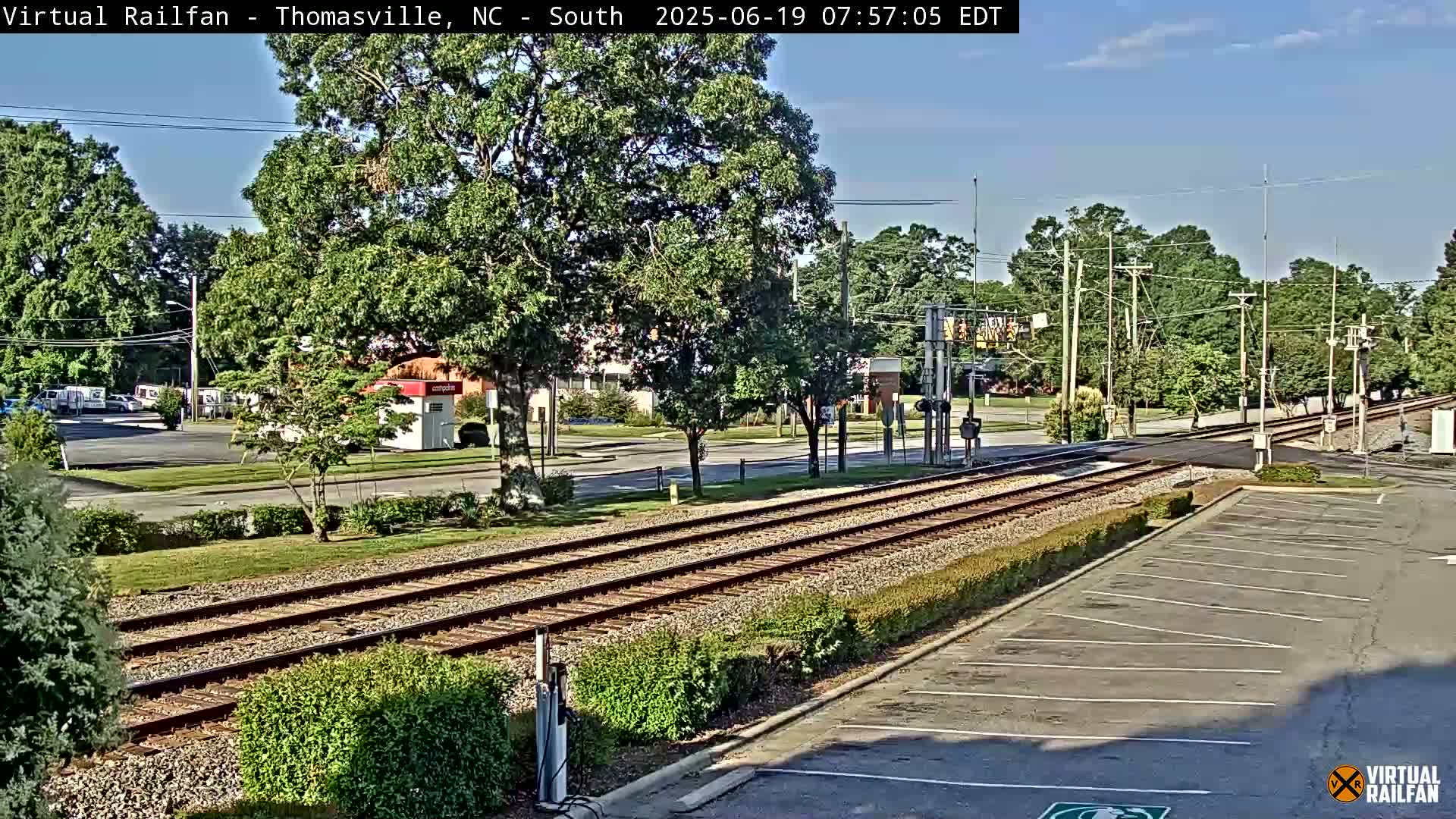 A sunny view of railroad tracks intersecting a road, surrounded by trees and a parking lot.