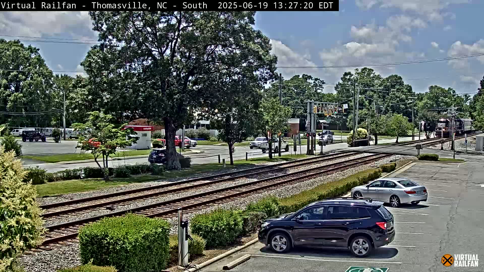 A train passes a road crossing under a partly cloudy sky, with several cars parked in a lot nearby.