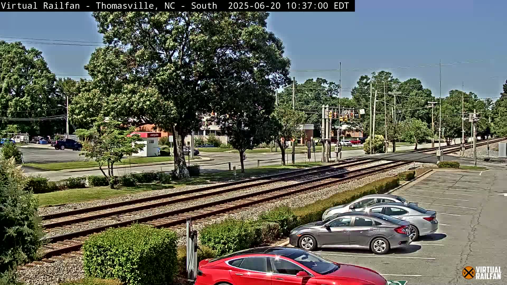 A sunny day reveals railroad tracks intersecting a street with parked cars and a few buildings in the distance, surrounded by green trees and landscaping.
