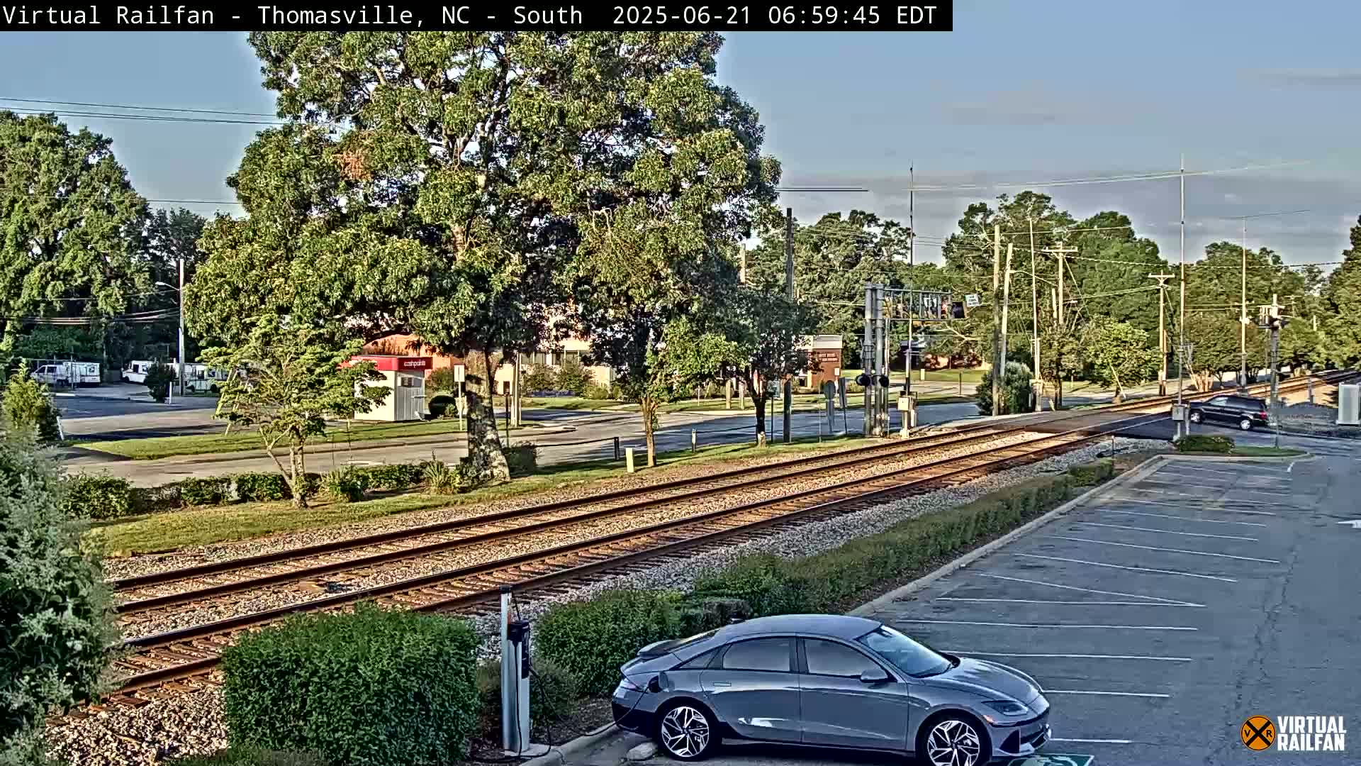 A gray car is parked near train tracks in a mostly sunny area, with some buildings and trees visible in the background.