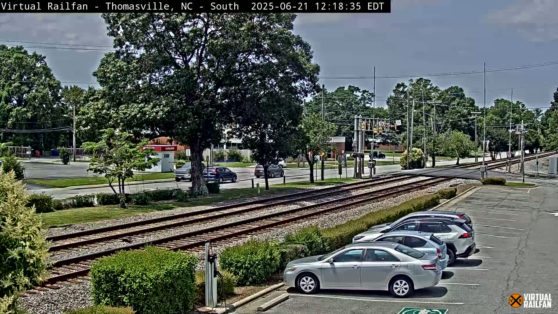 A sunny view of a parking lot next to train tracks, with several cars parked and a few cars driving on a nearby road lined with trees.
