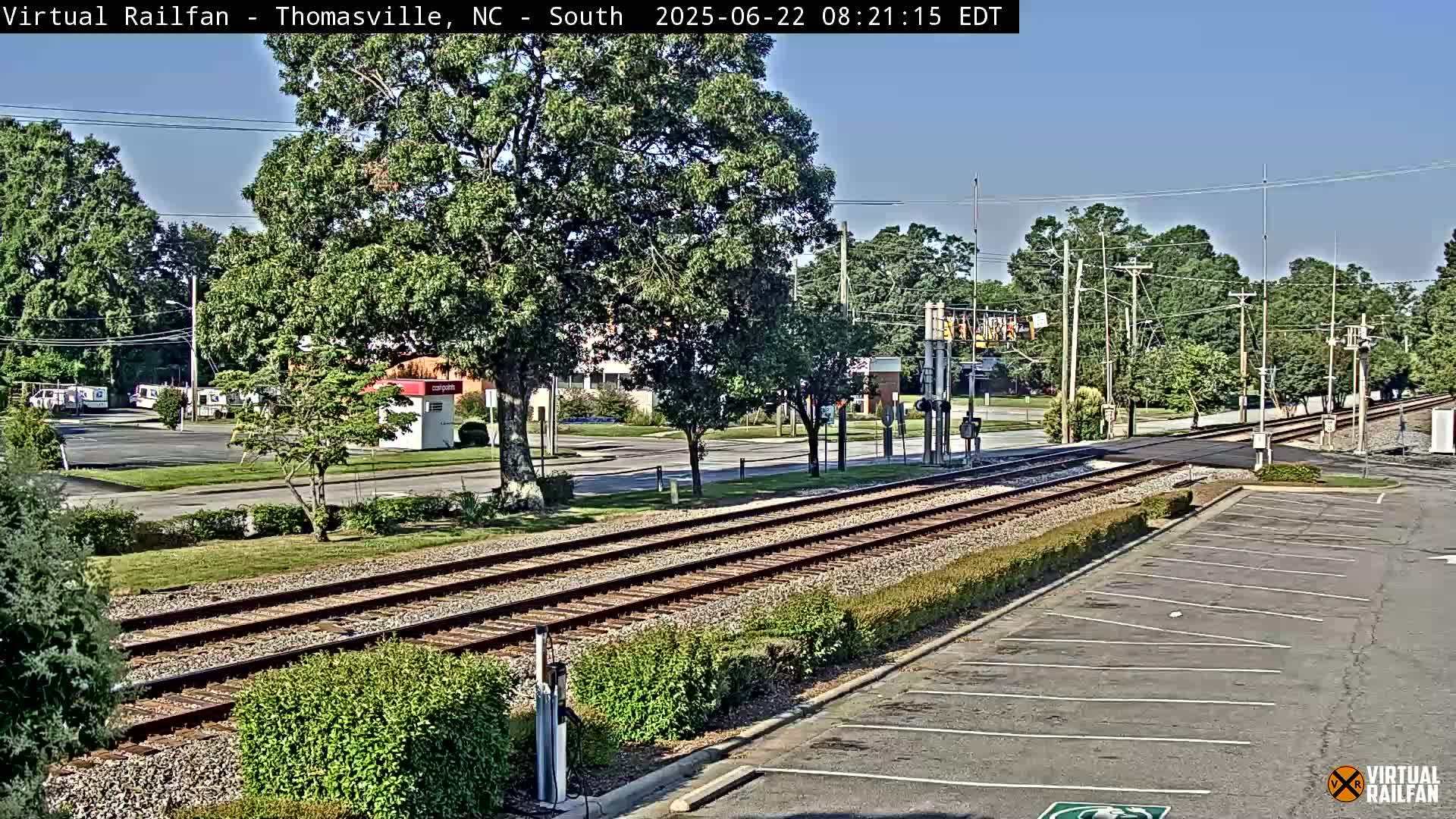 A sunny day reveals railroad tracks crossing a road in a suburban area with lush green trees and a mostly empty parking lot.