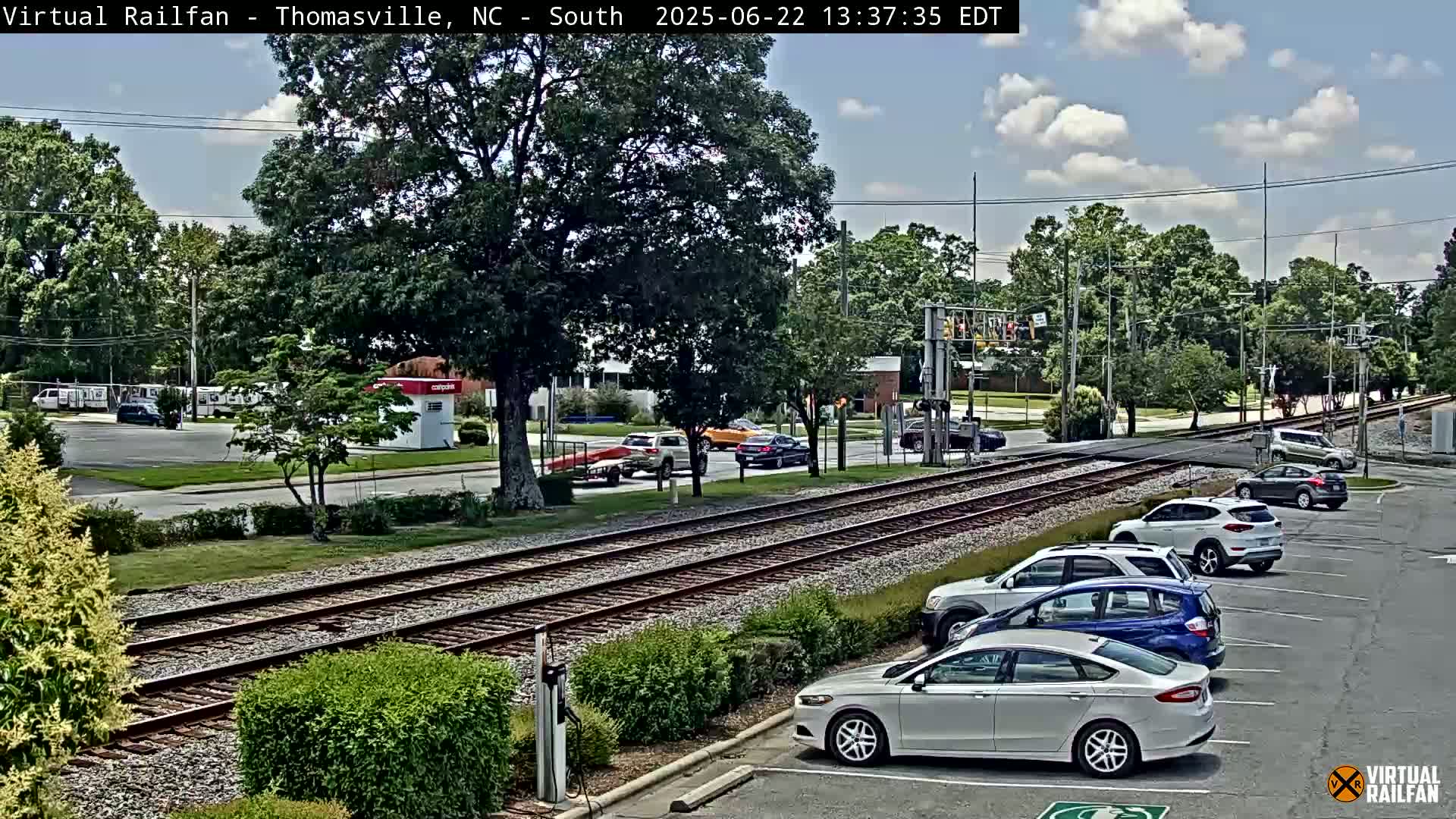 Under a partly cloudy sky, railroad tracks run parallel to a street with cars parked alongside, and several vehicles are visible driving on the road and crossing the tracks.