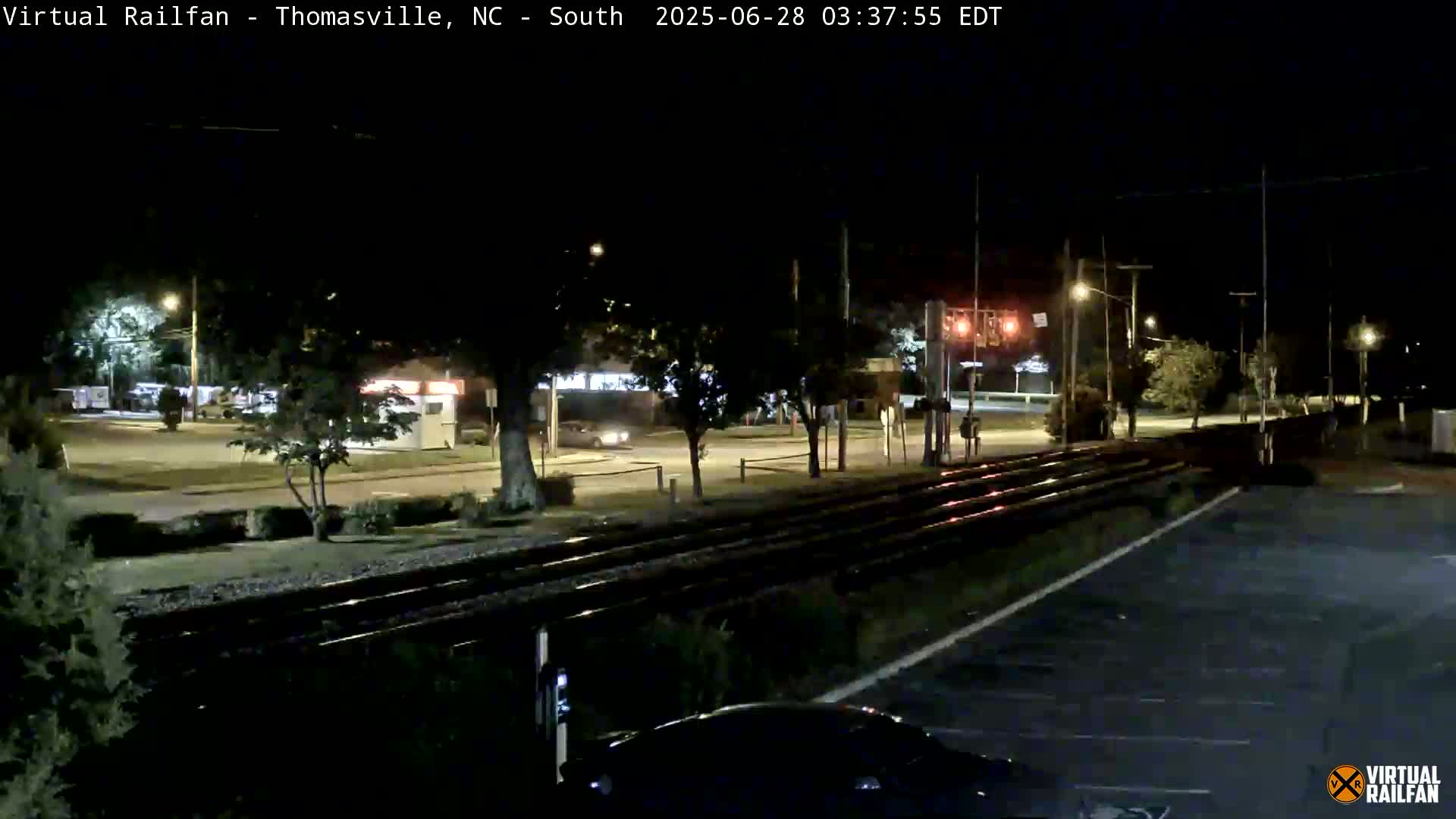 A nighttime view of railroad tracks intersecting a road, with cars visible on the road and sparse streetlights illuminating the scene.