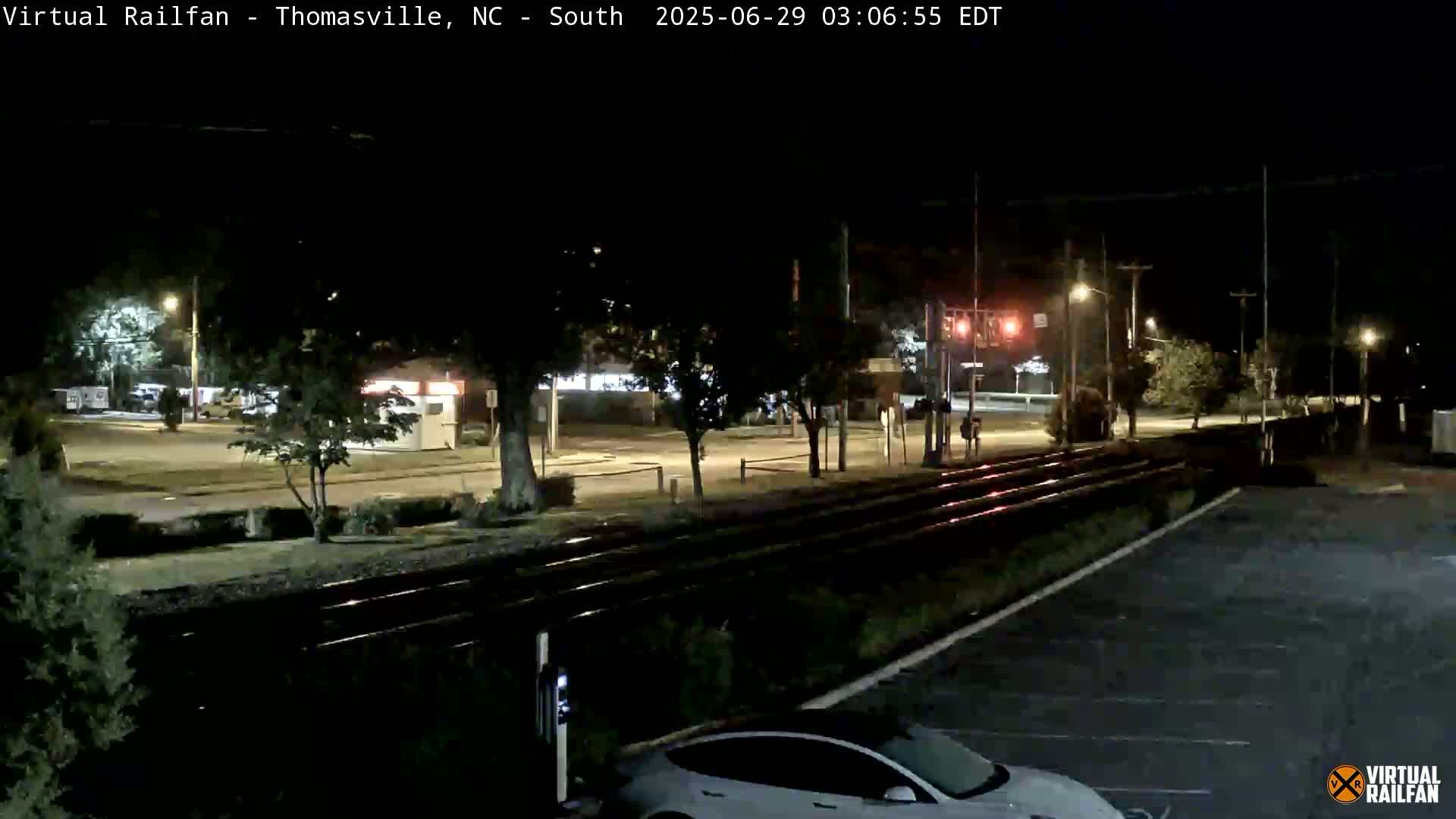 A nighttime view of railroad tracks and a parking lot, with a car parked near the tracks and streetlights illuminating the surrounding area.