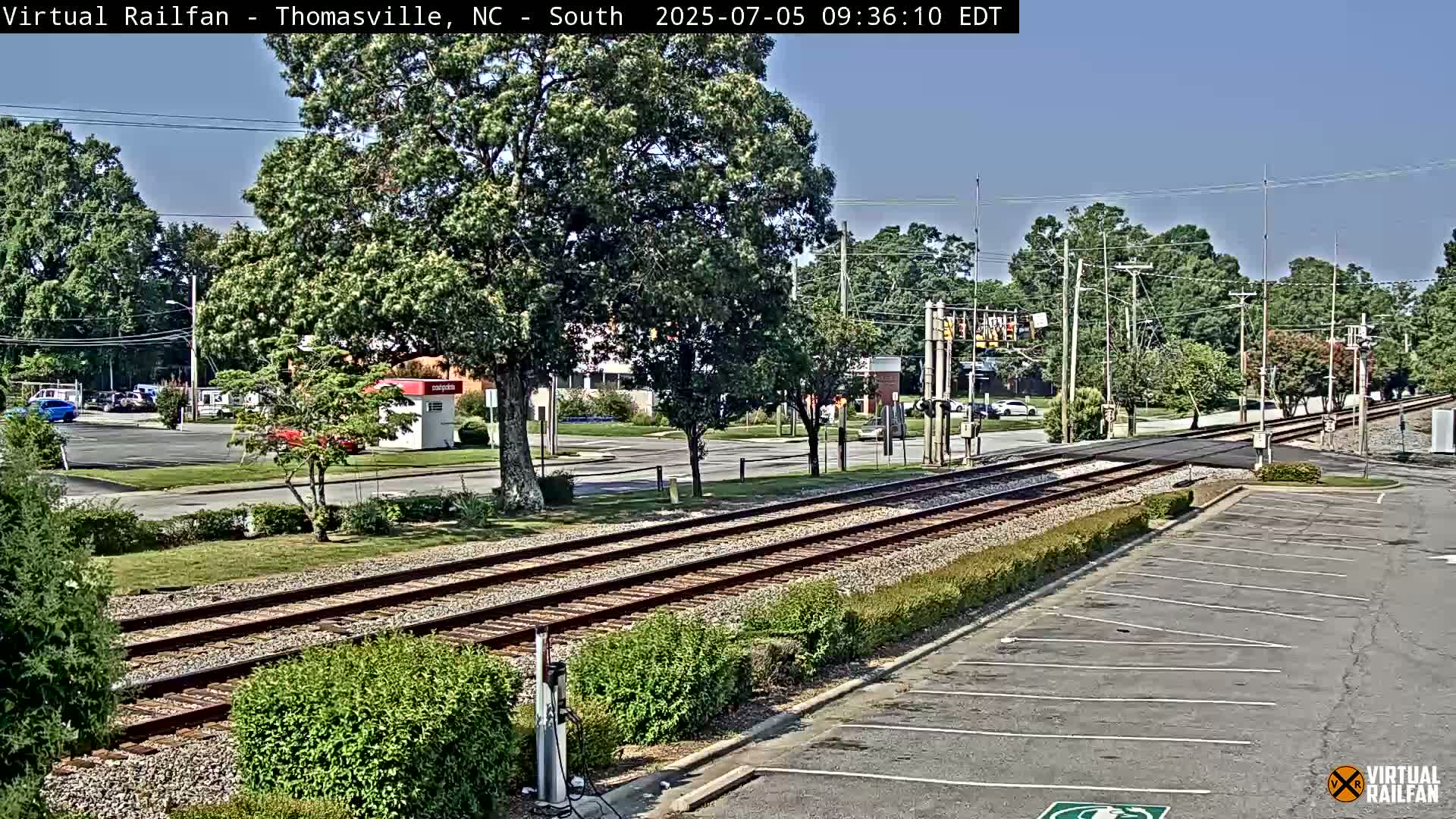 A sunny day view of railroad tracks crossing a road, surrounded by trees, shrubs, and a mostly empty parking lot.