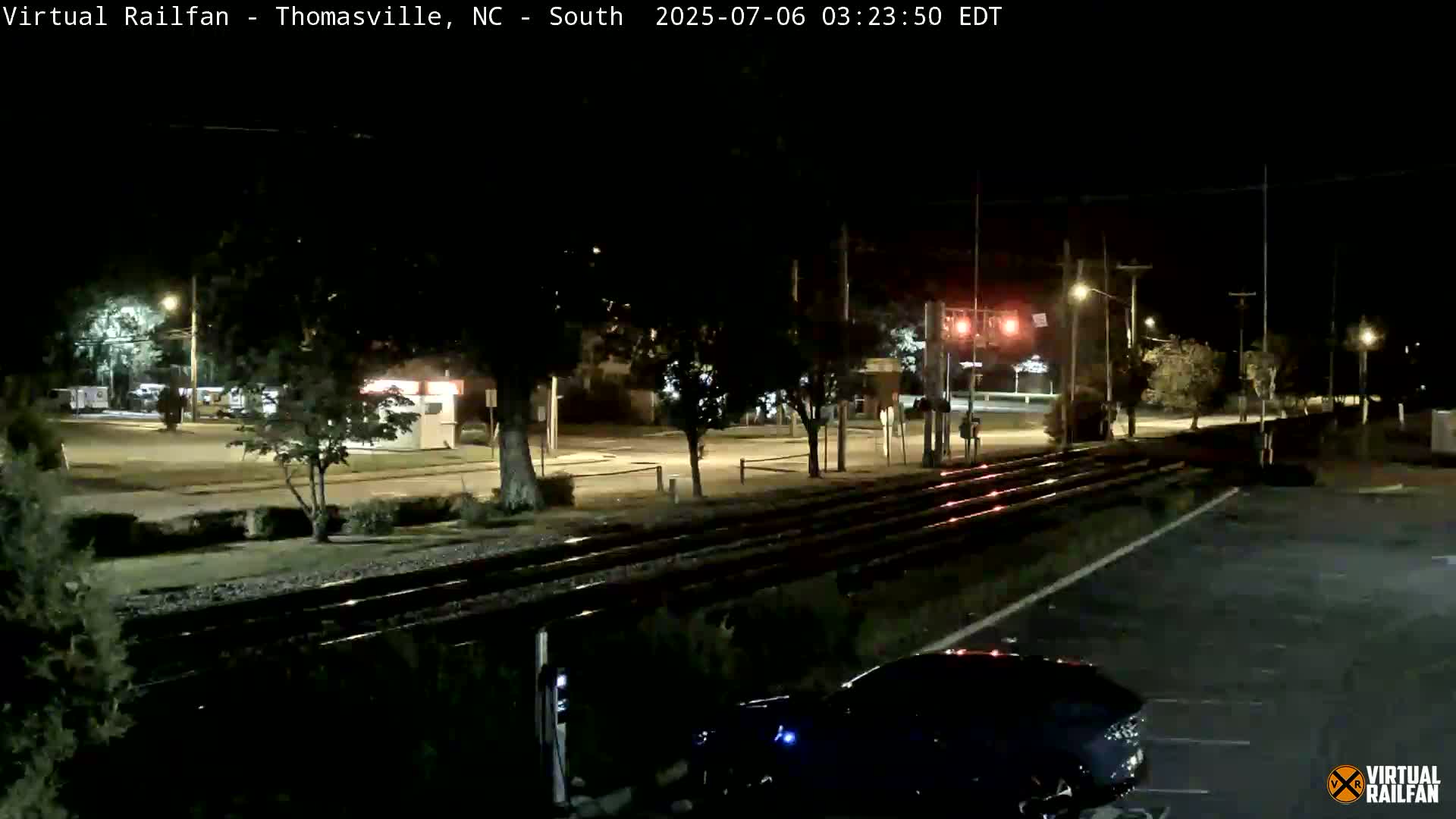 A nighttime view of railroad tracks crossing a street, with a car parked nearby under dark, clear skies.