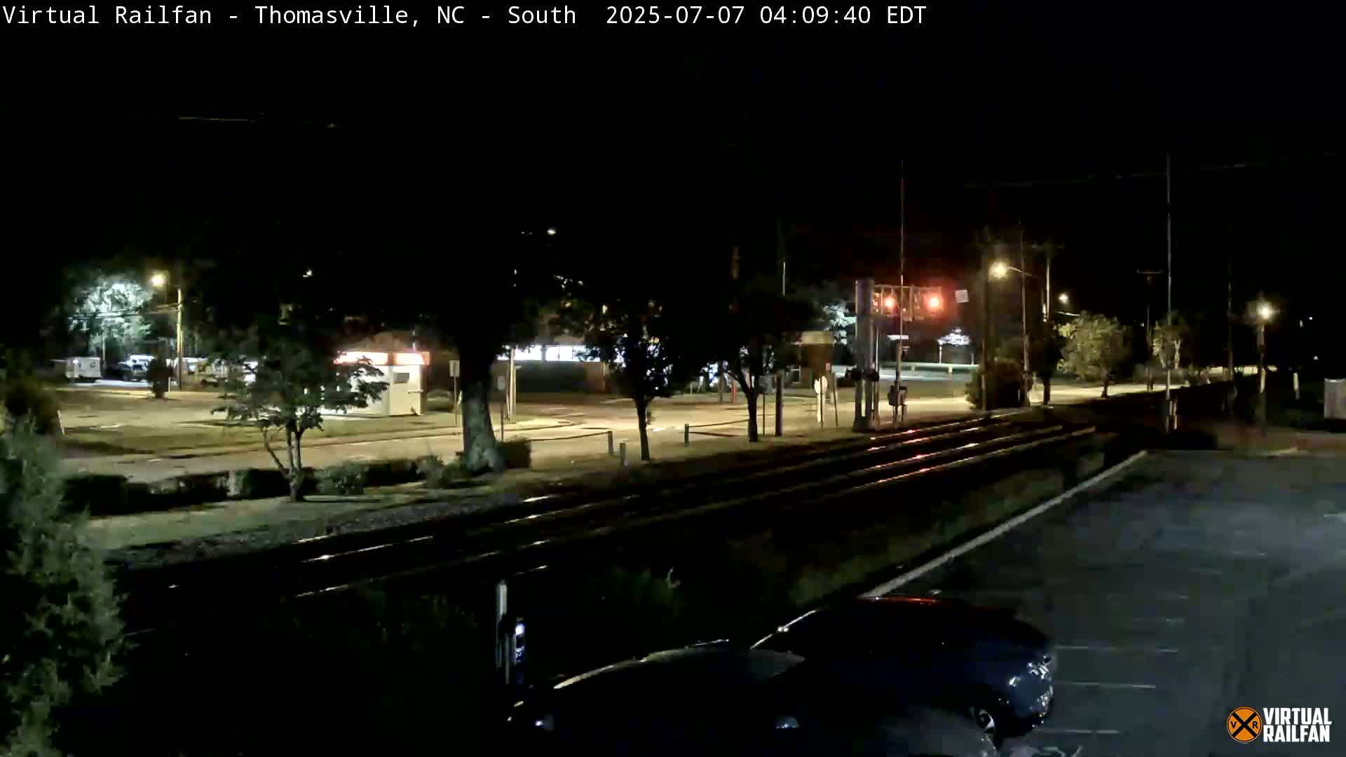A nighttime view of a train track intersecting a road, with cars parked in a lot nearby and streetlights illuminating the scene.