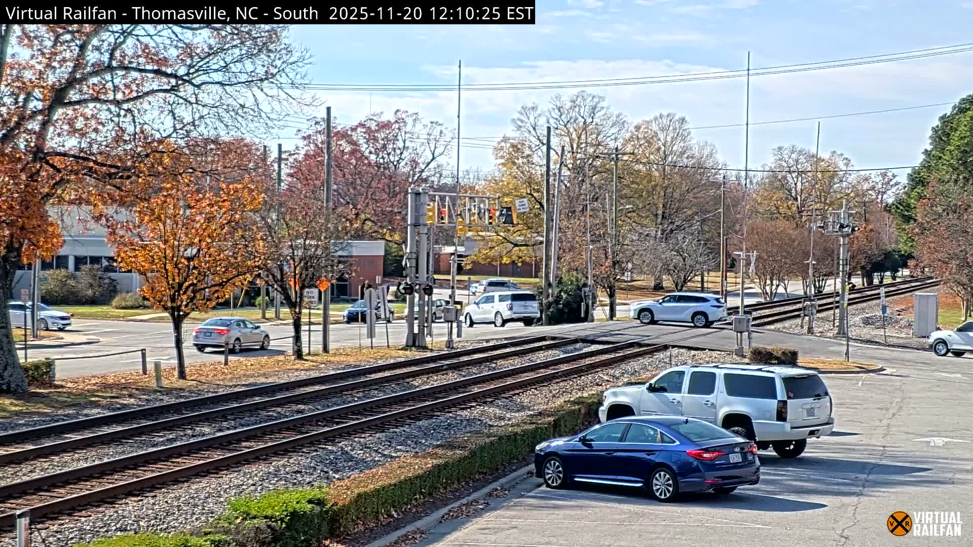 A bright, clear autumn day reveals a town scene with two railroad tracks running through, a road with several cars including a blue sedan and a white SUV, and many trees displaying vibrant fall foliage.