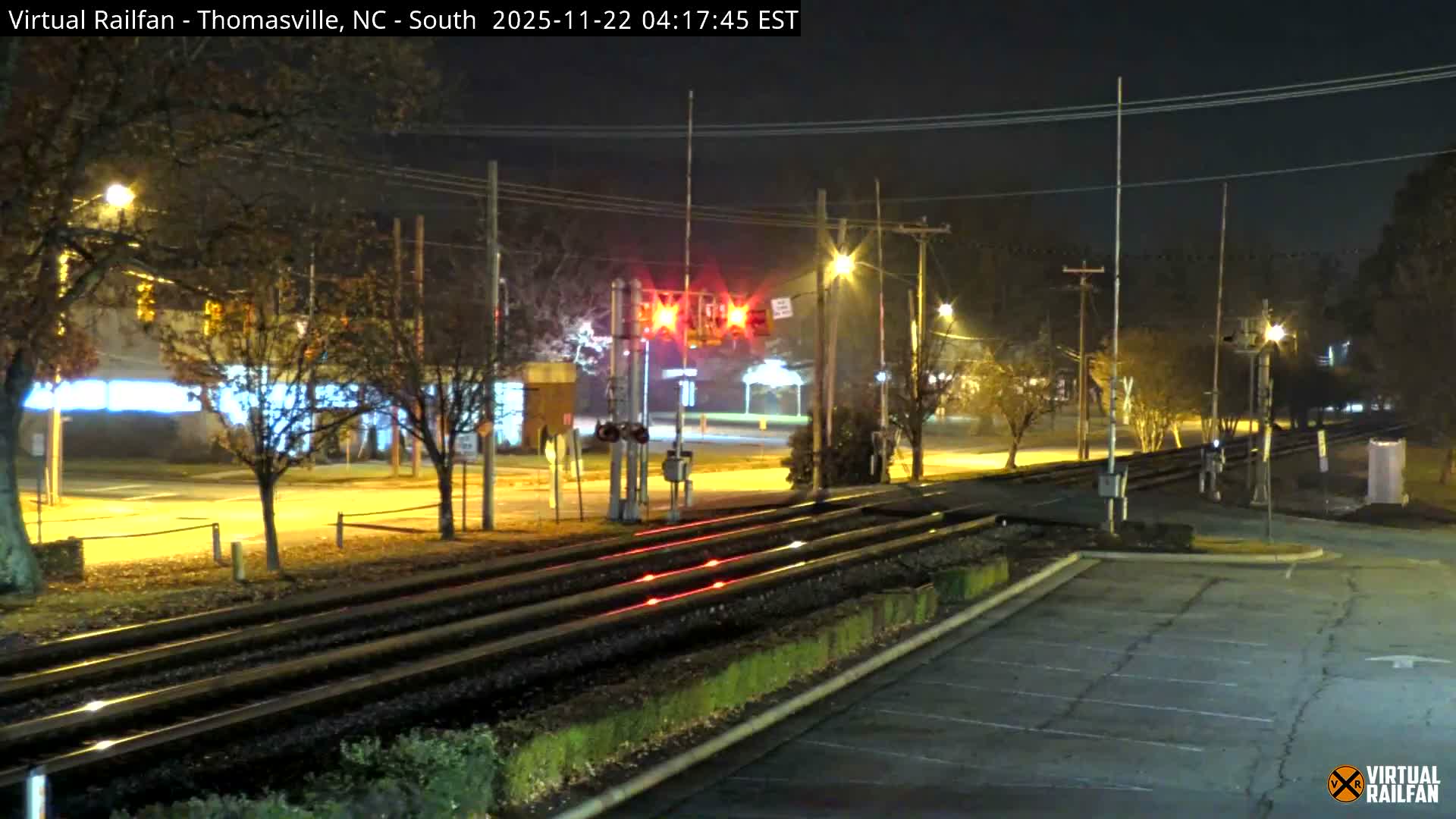 An active railroad crossing with flashing red warning lights and upright gate arms spans multiple tracks, illuminated by streetlights at night under a clear, calm sky.