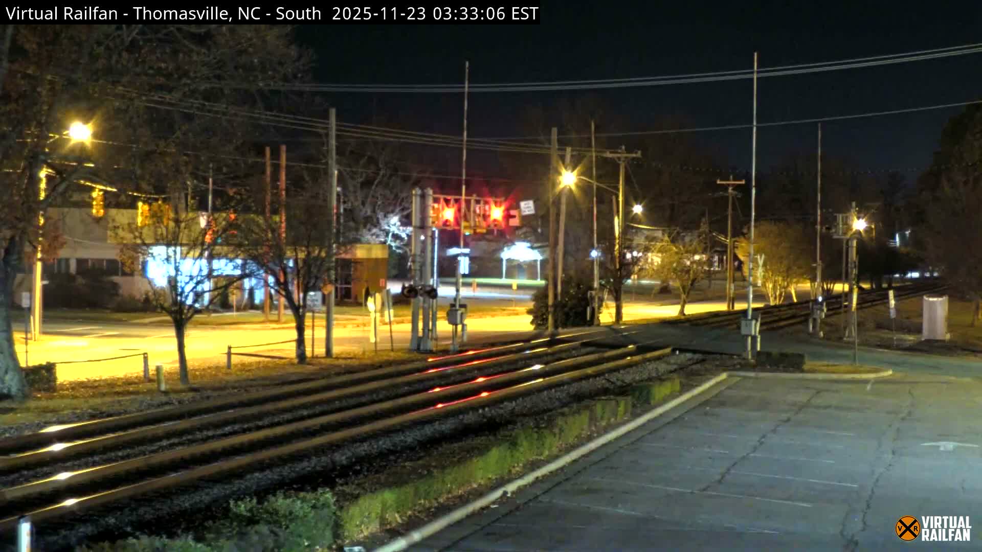 A clear night reveals an active multi-track railroad crossing illuminated by streetlights, with red crossing signals and yellow traffic lights visible in the midground.