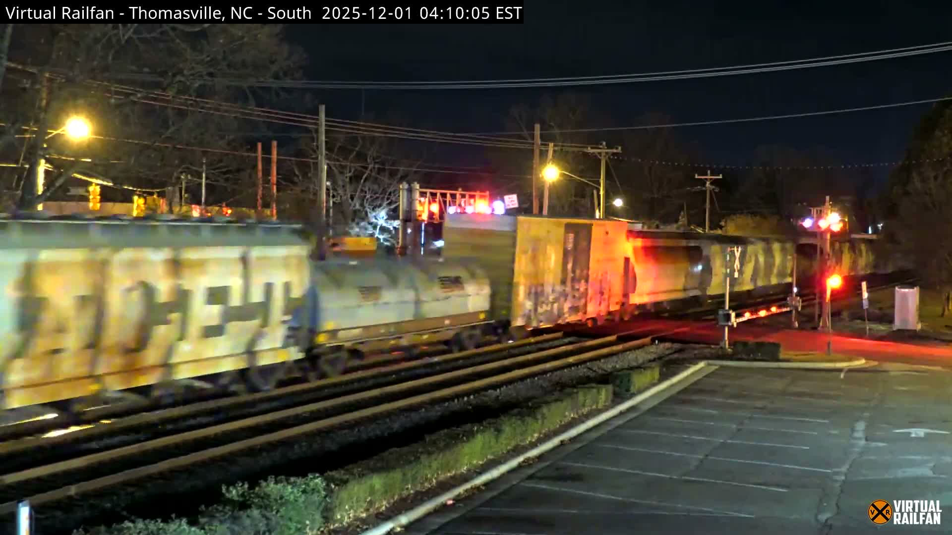A long freight train, illuminated by its own lights and flashing red crossing signals, is actively passing through a multi-track railway crossing on a clear night.