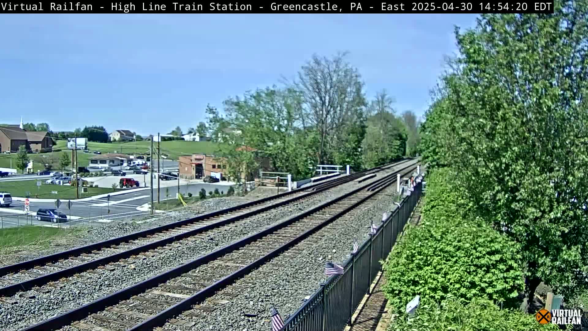 A sunny view of railroad tracks extending into the distance, passing through a small town with a mix of buildings and greenery.