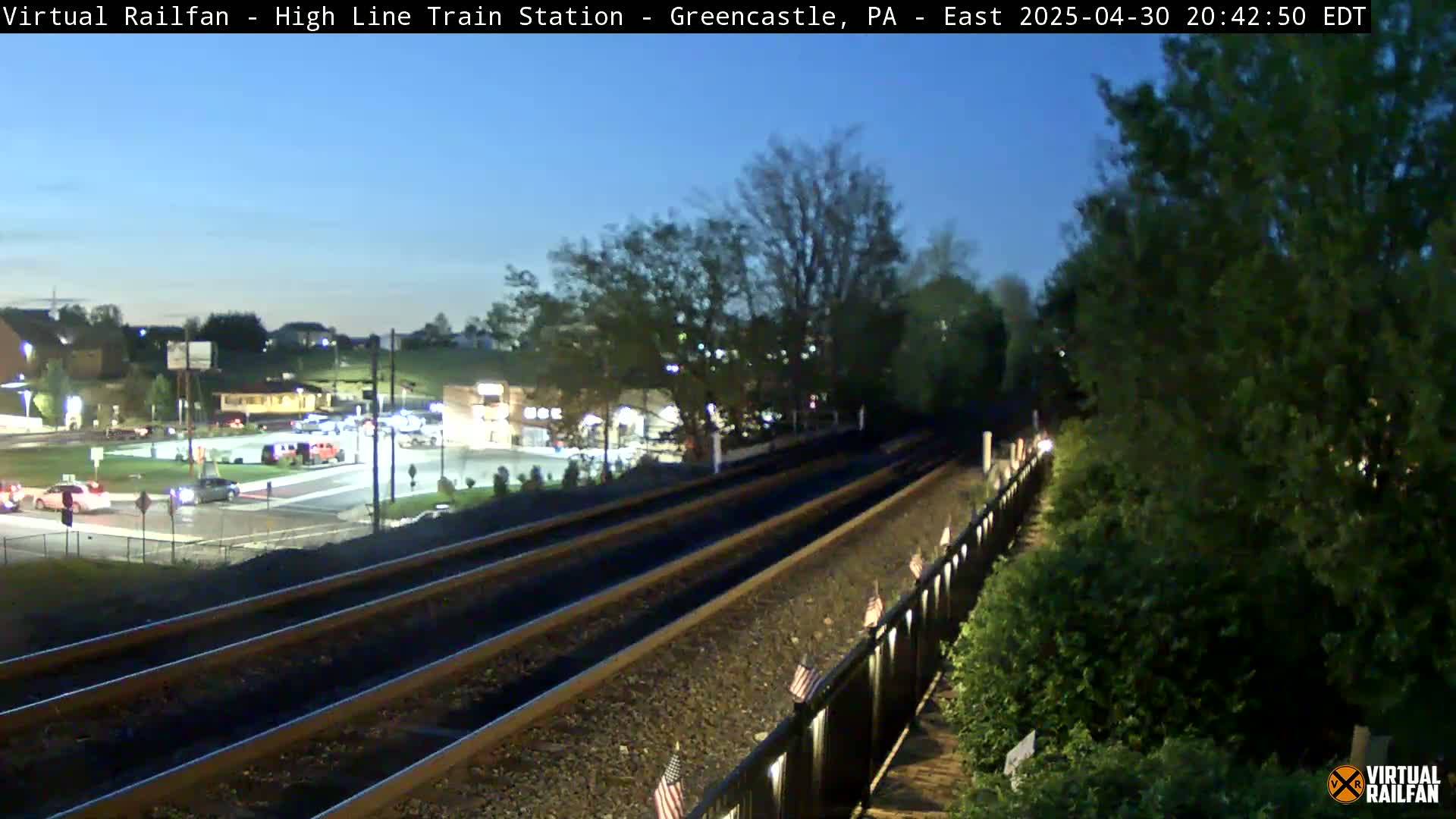 A twilight view of train tracks extending from a dimly lit station area, past a fence adorned with small flags, towards a tree line.