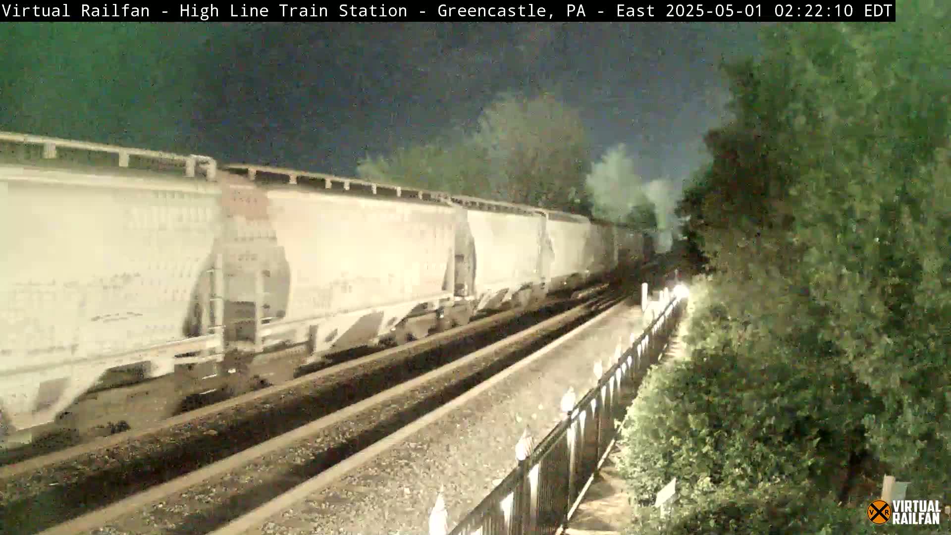 At night, a long freight train of white covered hopper cars passes by on a double track railroad line next to a dark green shrubbery.