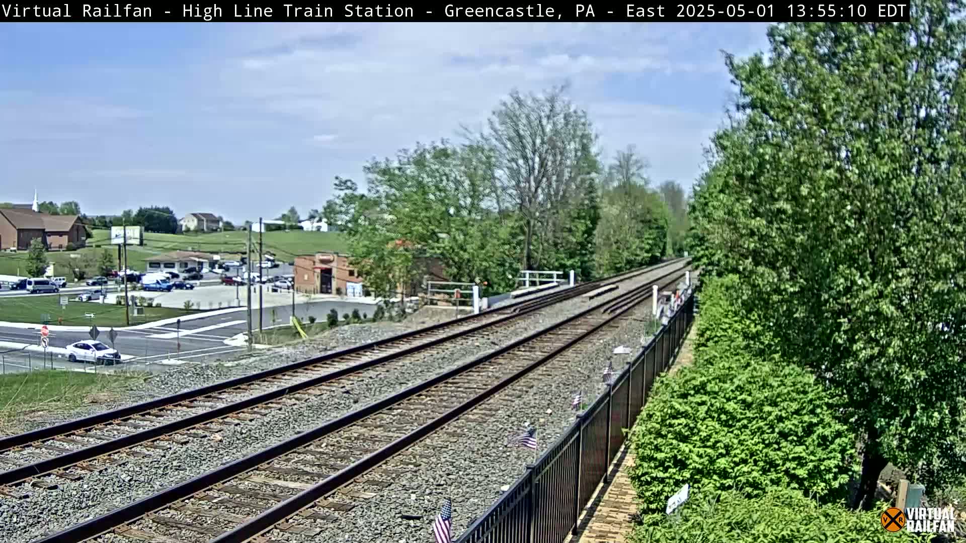 A view of train tracks extending into the distance on a partly sunny day, with green trees and bushes lining the sides, and a small town visible in the background.