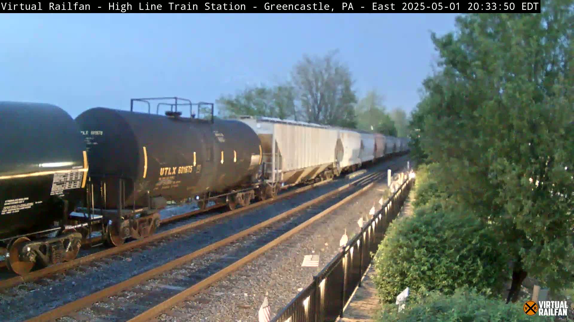 A long freight train, consisting of several dark tank cars and light-colored boxcars, passes by under a twilight sky.