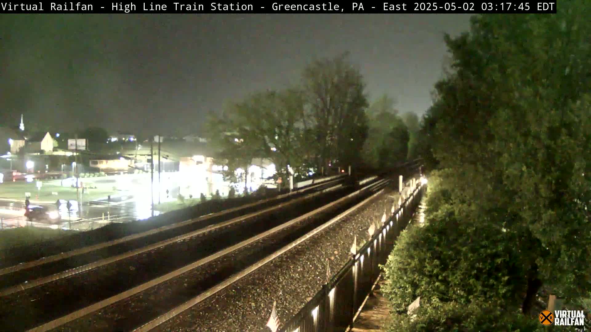 A nighttime view of a wet train track, with a town visible in the background under a dark, overcast sky.