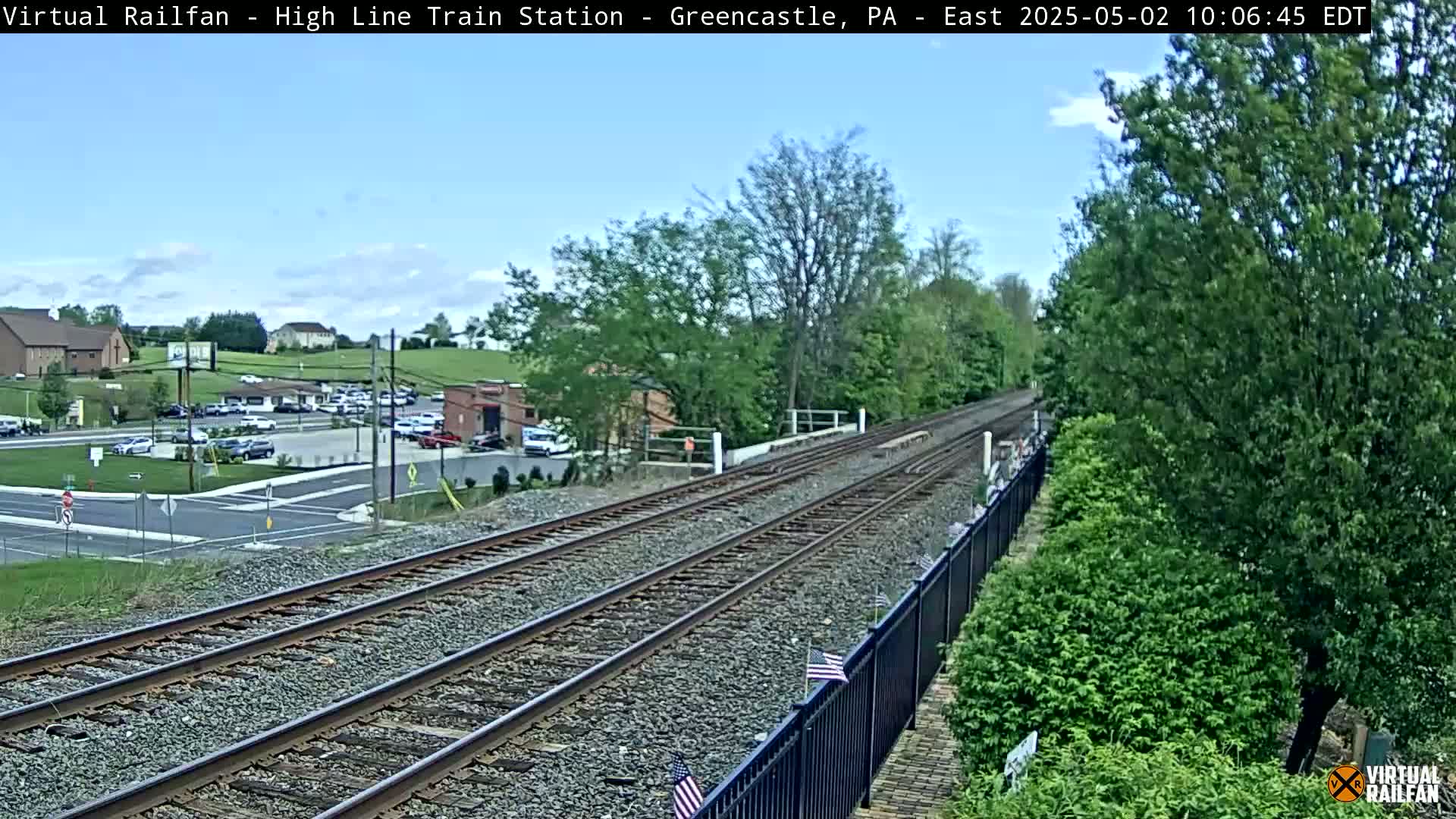 A view of a train track stretching into the distance on a partly sunny day, with green trees and a parking lot visible in the background.