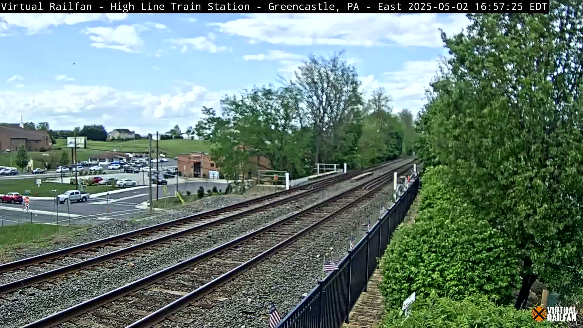 Under partly cloudy skies, a view of train tracks extends from a parking lot with several cars to a tree-lined area.