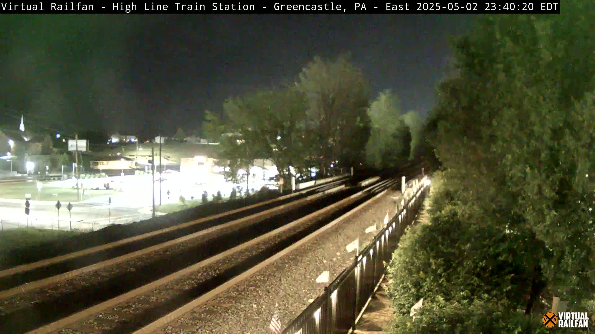 At night, three sets of train tracks extend into the distance, bordered by a fence and lush green vegetation, with a brightly lit intersection visible in the background.
