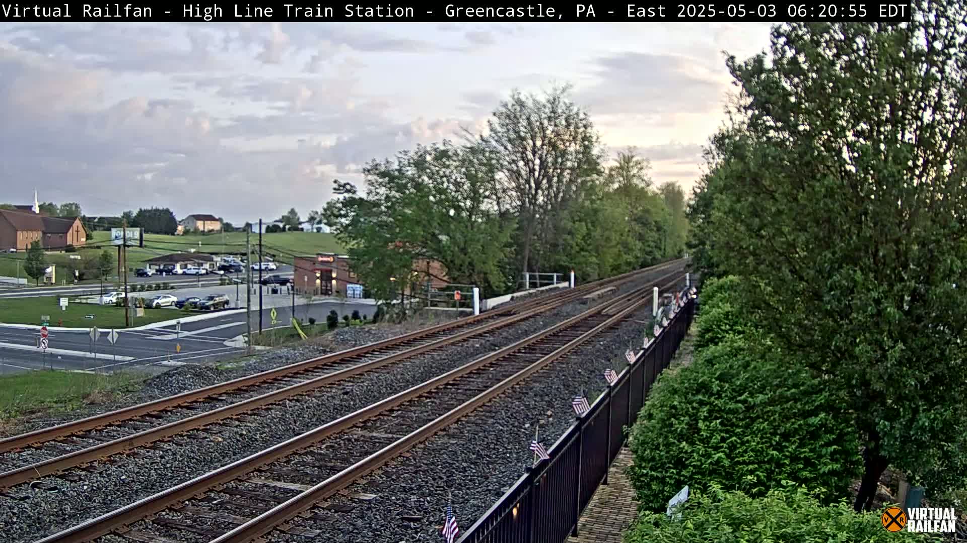 A high-angle, long shot shows a train track extending into the distance under a partly cloudy, twilight sky, with green trees and a small town visible in the background.